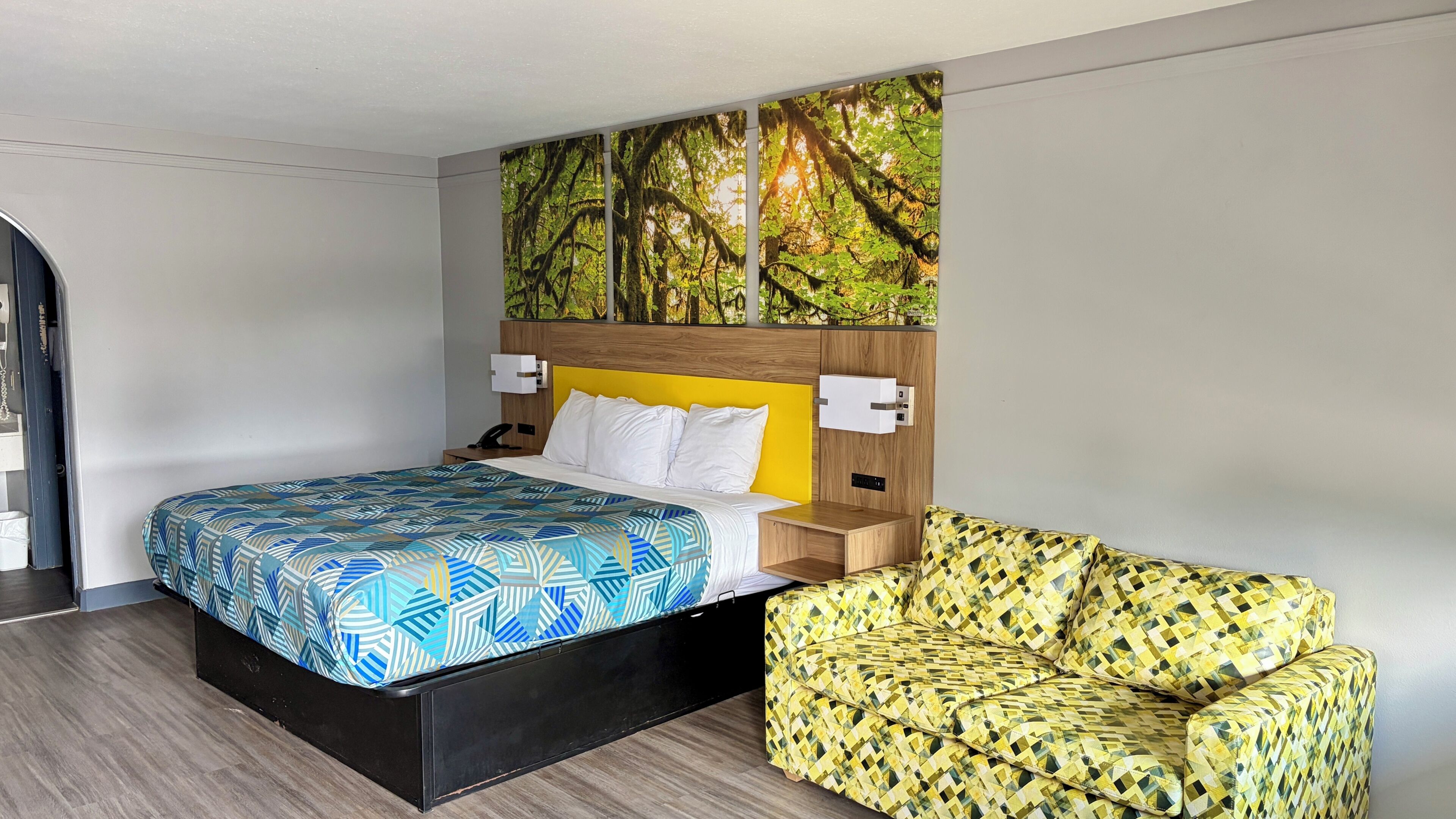 A hotel room with a bed, a nightstand, a chair, and a wall mural.