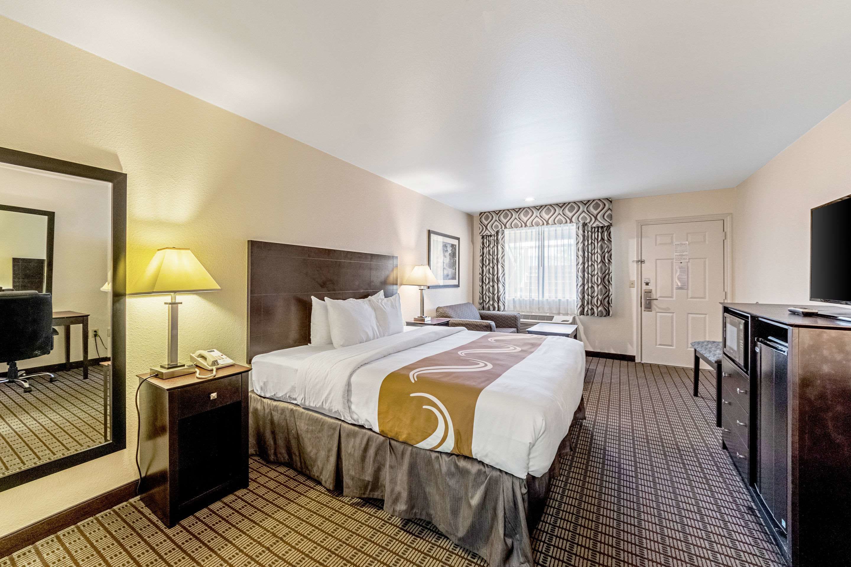 A hotel room with a large bed, a desk, a chair, a TV, a mirror, and a window with patterned curtains.