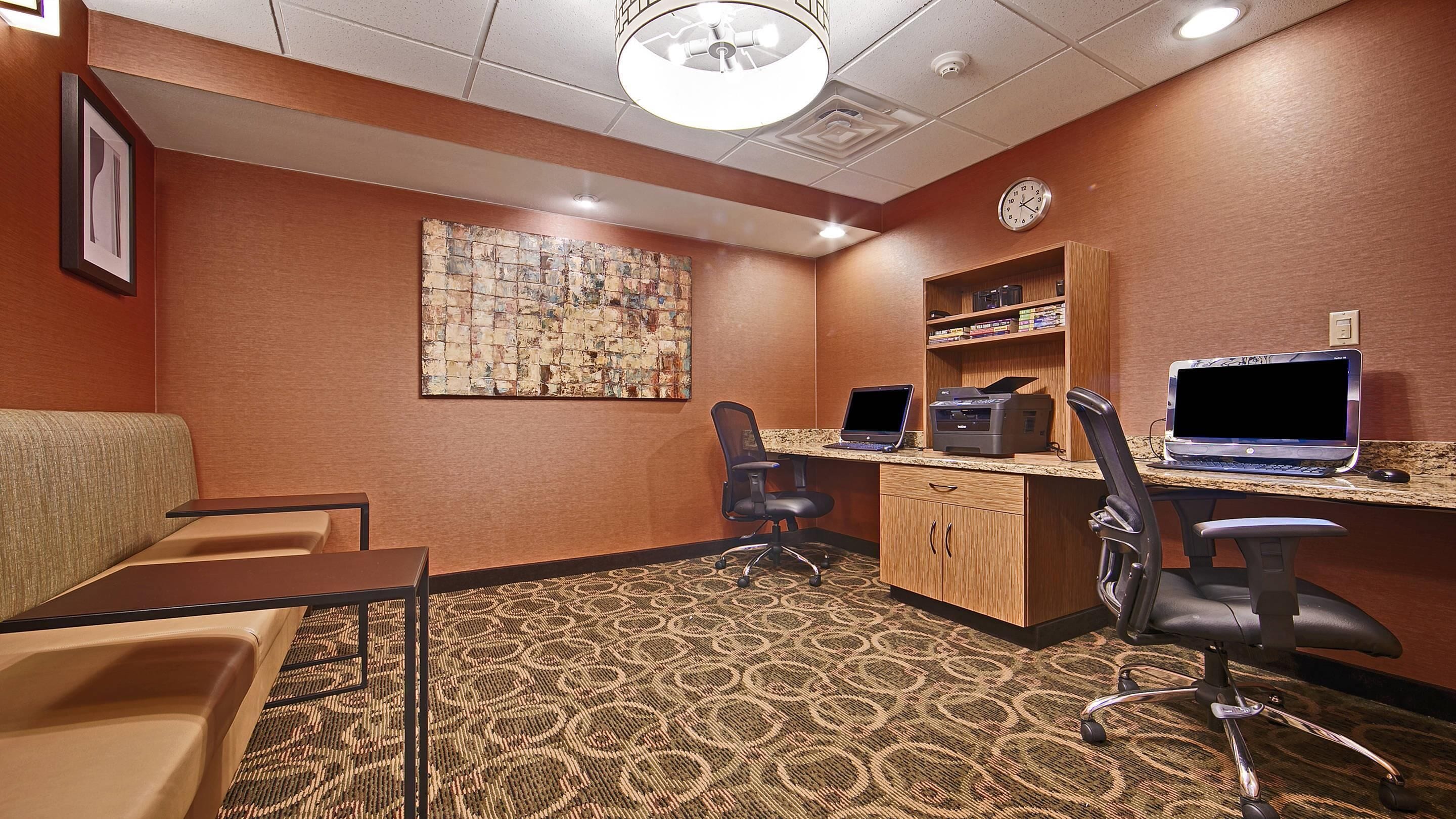 A conference room with a carpeted floor, a desk with a computer and printer, two office chairs, a sofa, and a clock on the wall.