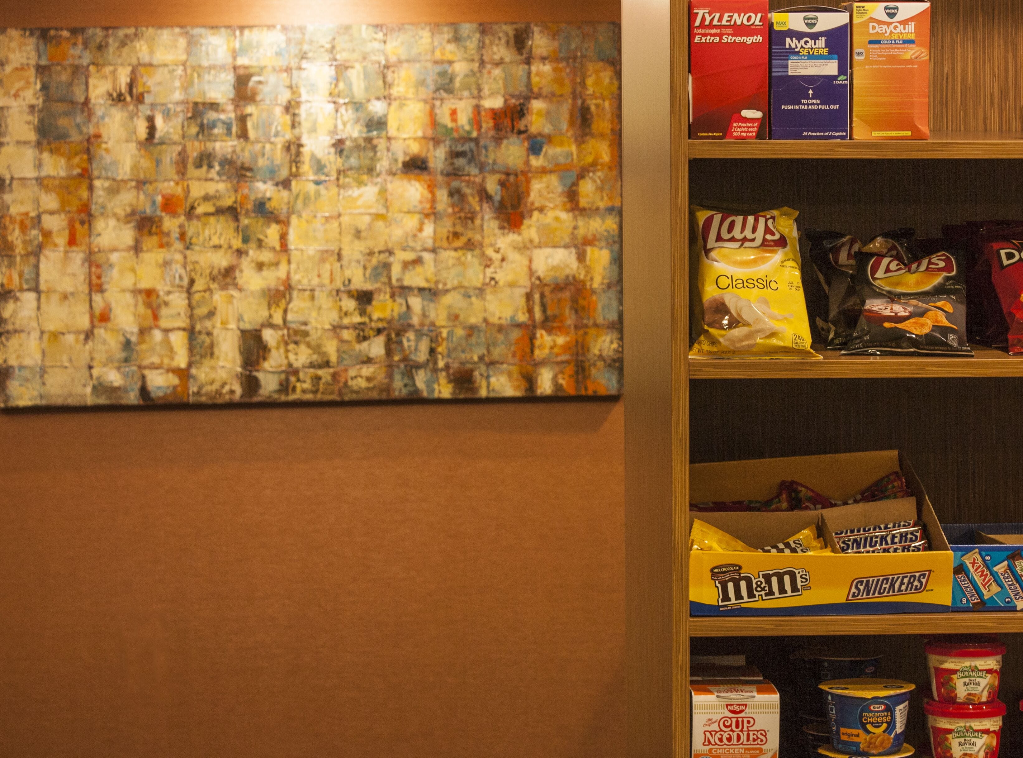 En væg med et farverigt abstrakt maleri, hylder med snacks og drikkevarer, samt en æske med M&Ms.