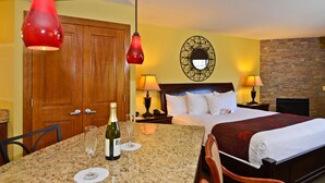 A hotel room with a bed, a dining table with a bottle of wine and glasses, a wooden door, and a wall with a decorative mirror.