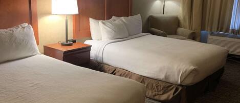 A hotel room with two beds, a nightstand, a lamp, a chair, and a window with curtains.