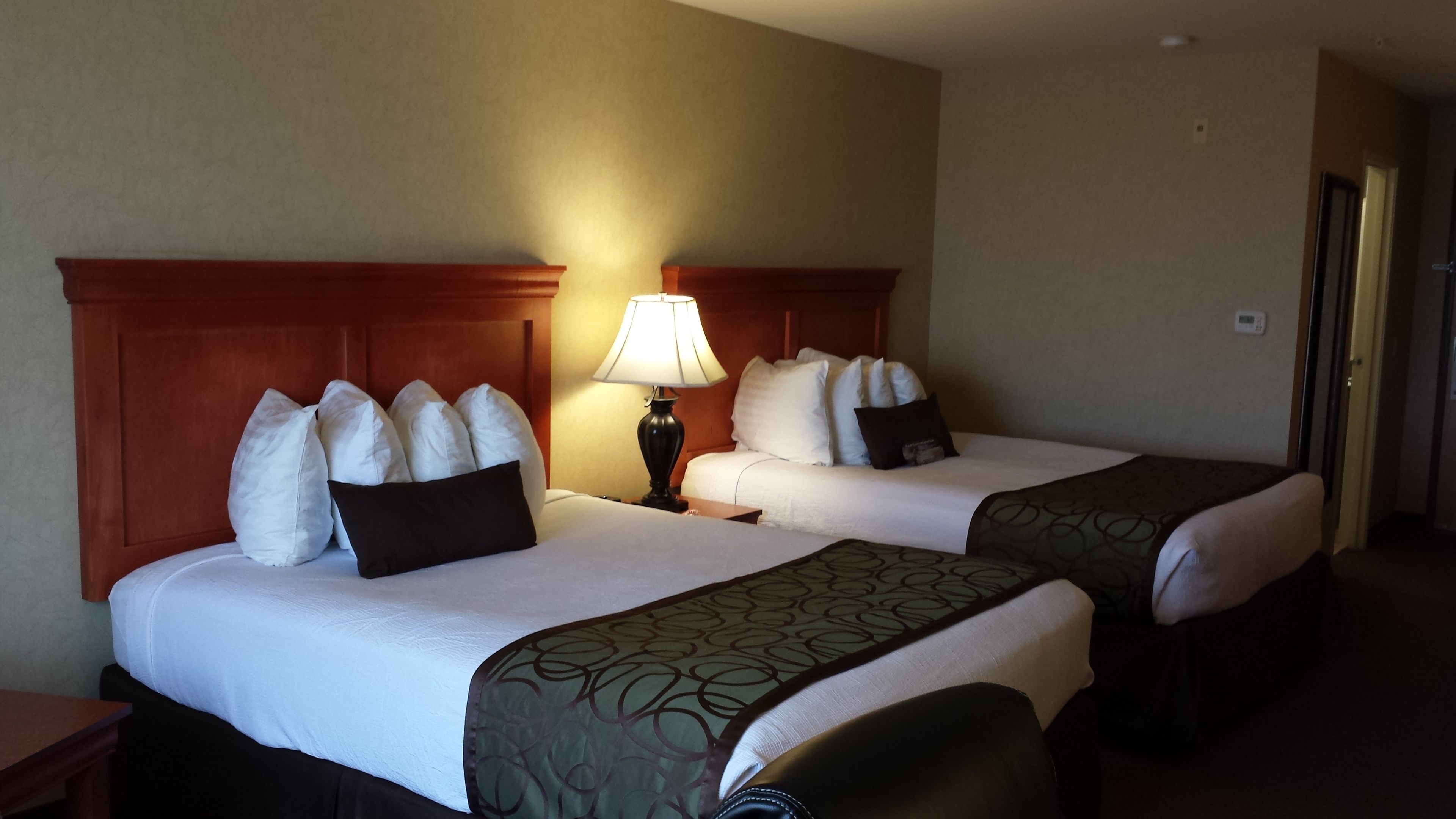 A hotel room with two beds, each with white bedding and multiple pillows. There is a wooden headboard, a bedside table with a lamp, and a door to the right.