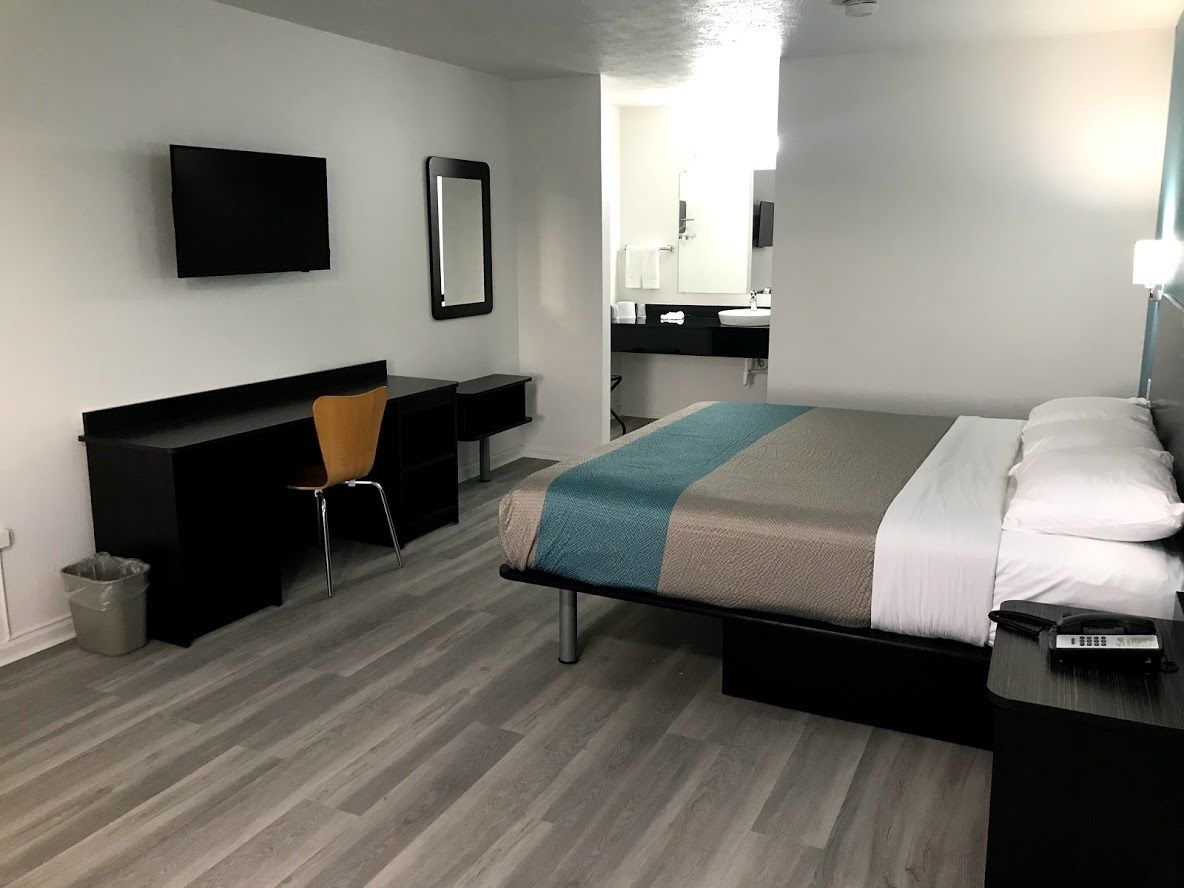 A modern hotel room with a bed, a desk, a chair, a TV, and a bathroom visible through an open door.