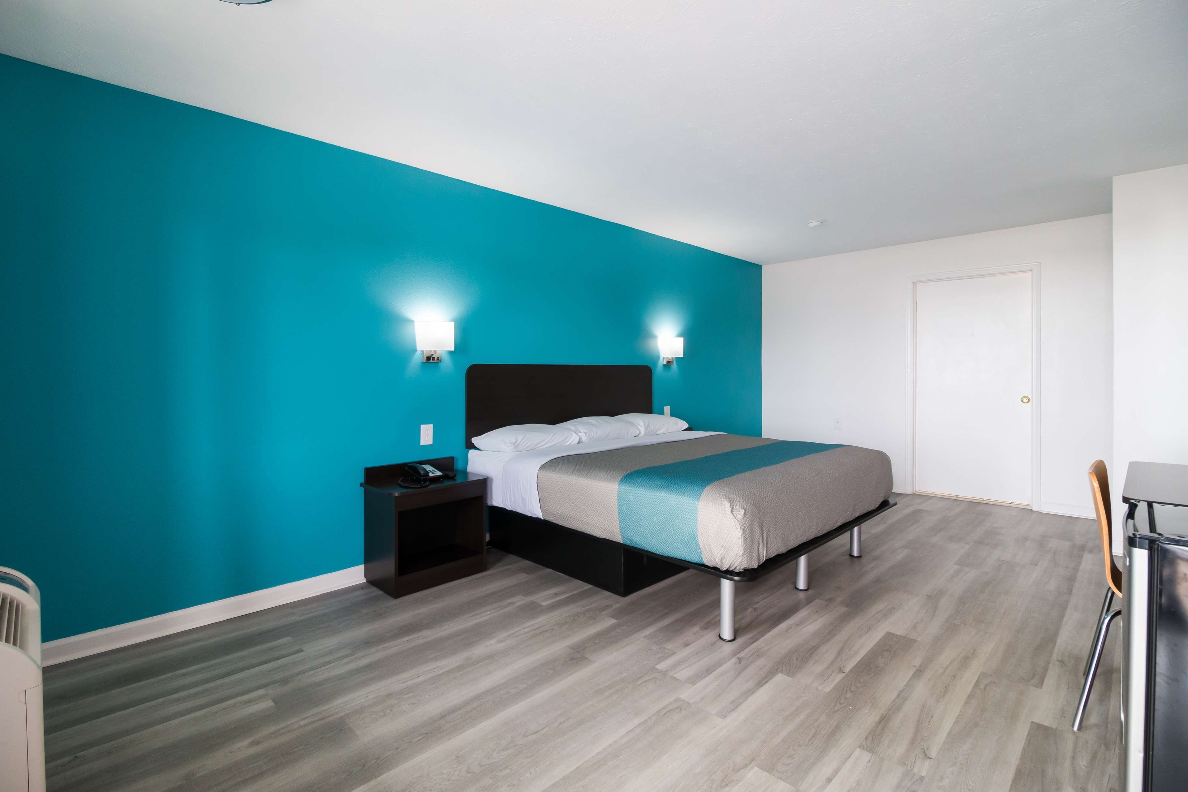 Una moderna camera d'albergo con una parete verde acqua, un letto con copripiumino grigio e verde acqua, un comodino e una scrivania con una sedia.