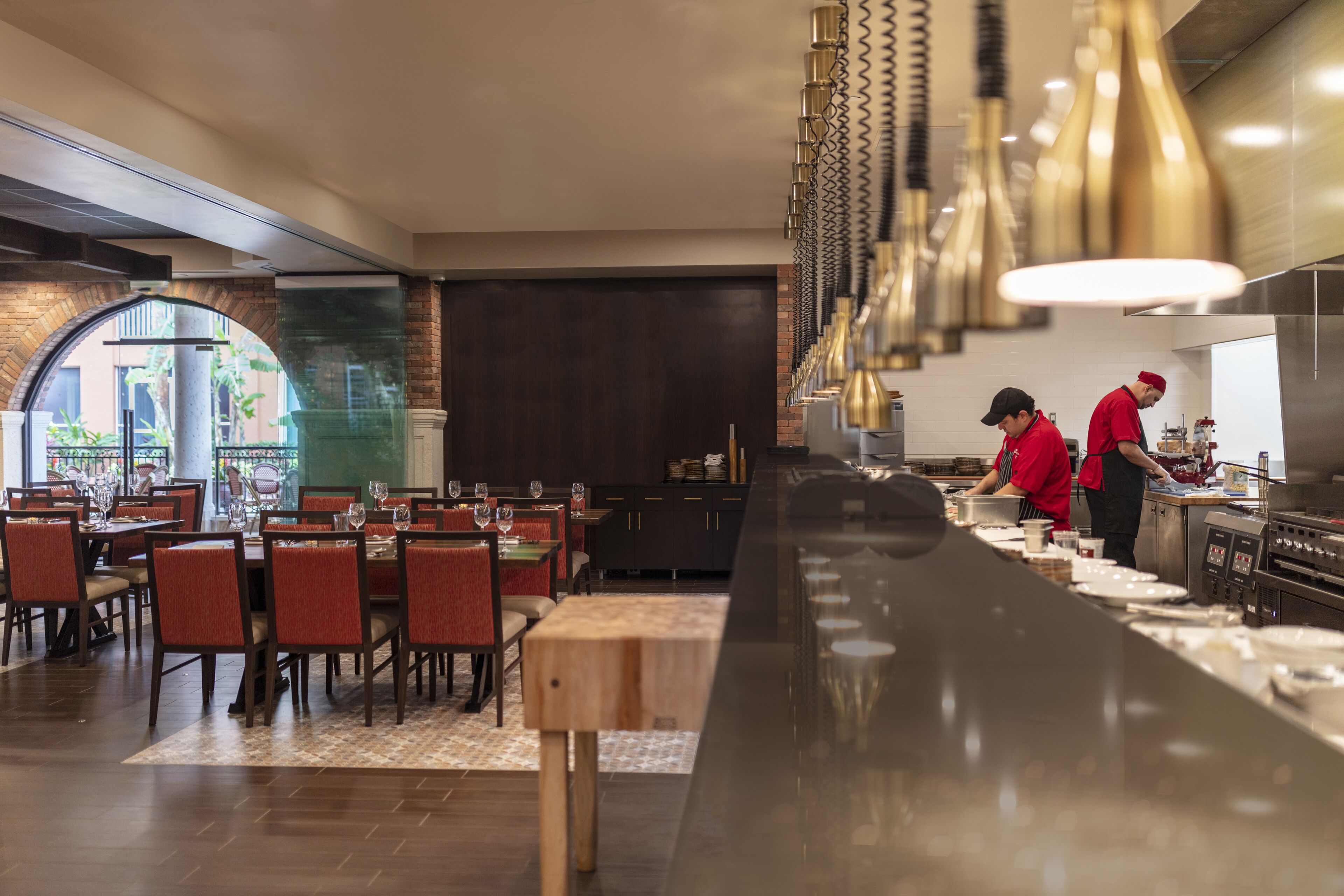 Ein Restaurant mit einer langen Theke, Esstischen und Stühlen. Zwei Kellner tragen rote Uniformen.