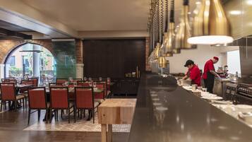 A restaurant with a long counter, dining tables, and chairs. There are two waiters in red uniforms.