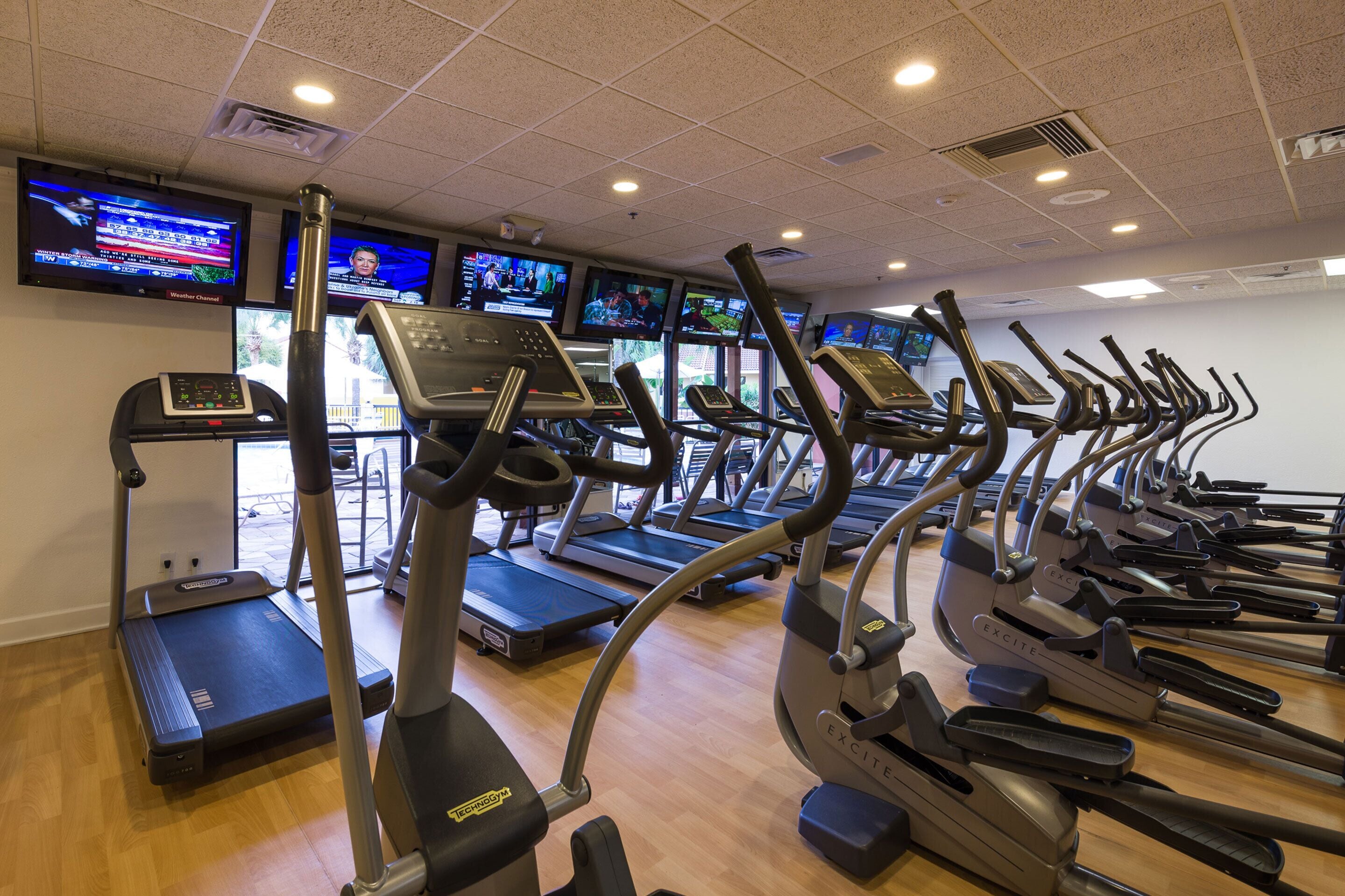A gym with multiple treadmills and elliptical machines, a TV wall displaying news channels, and a wooden floor.