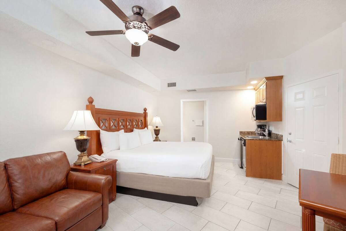 A hotel room with a bed, a brown leather sofa, a desk, a ceiling fan, and a kitchenette.