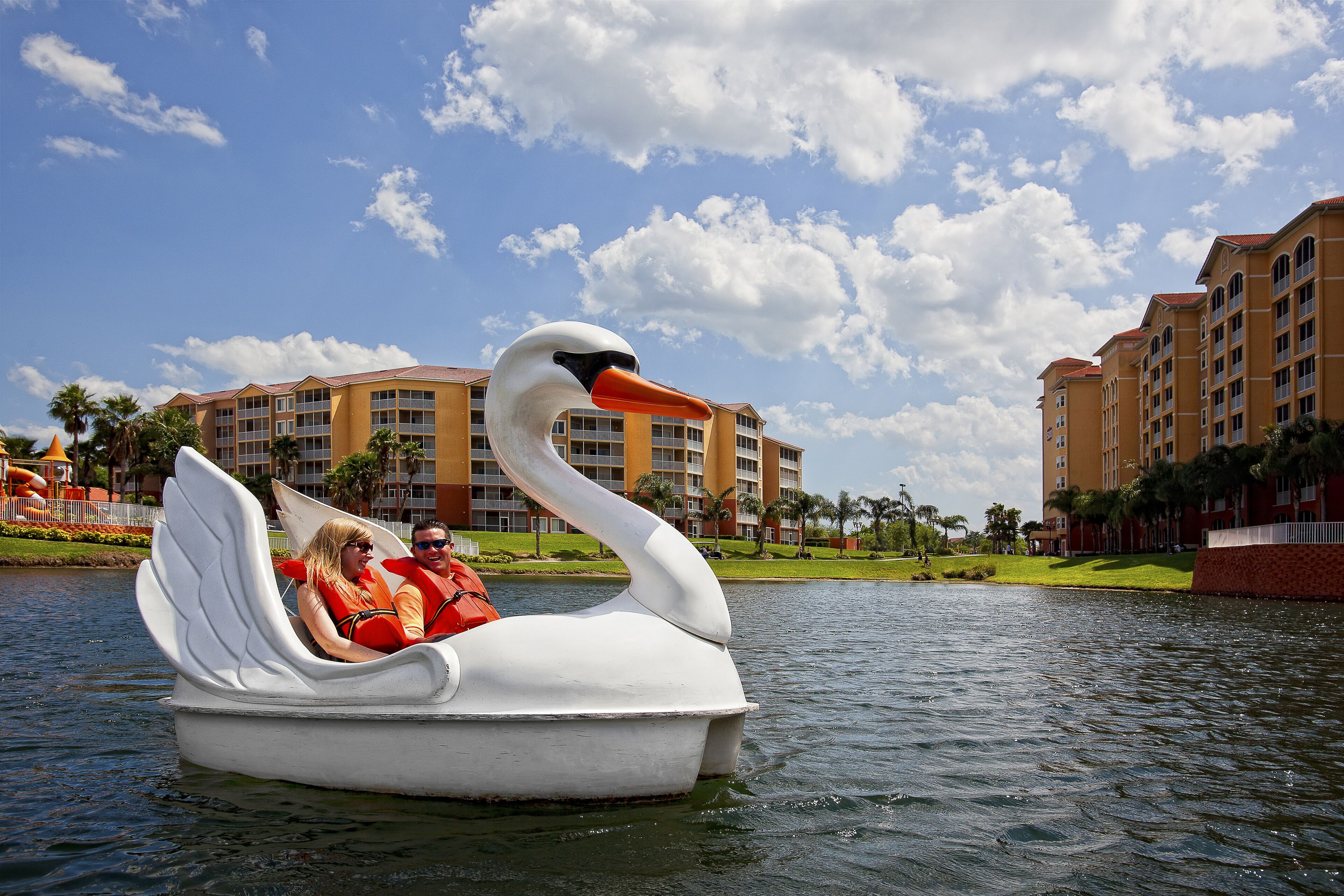 A swan-shaped boat with two people on it, floating on water with hotel buildings in the background.