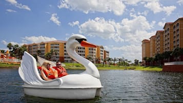 A swan-shaped boat with two people on it, floating on water with hotel buildings in the background.
