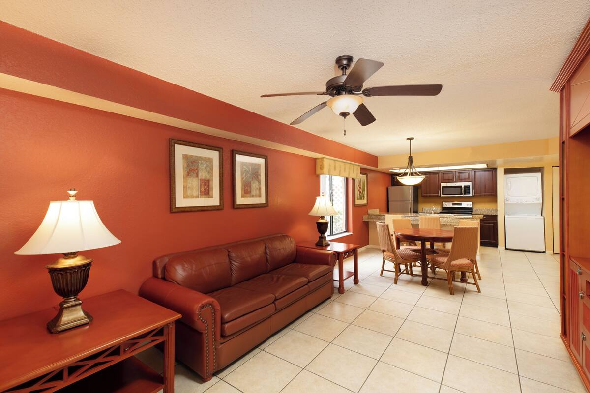 A living room with a brown leather sofa, a wooden coffee table, and a dining area with a wooden table and chairs. There is a ceiling fan, a kitchen with a microwave and refrigerator, and framed pictures on the wall.