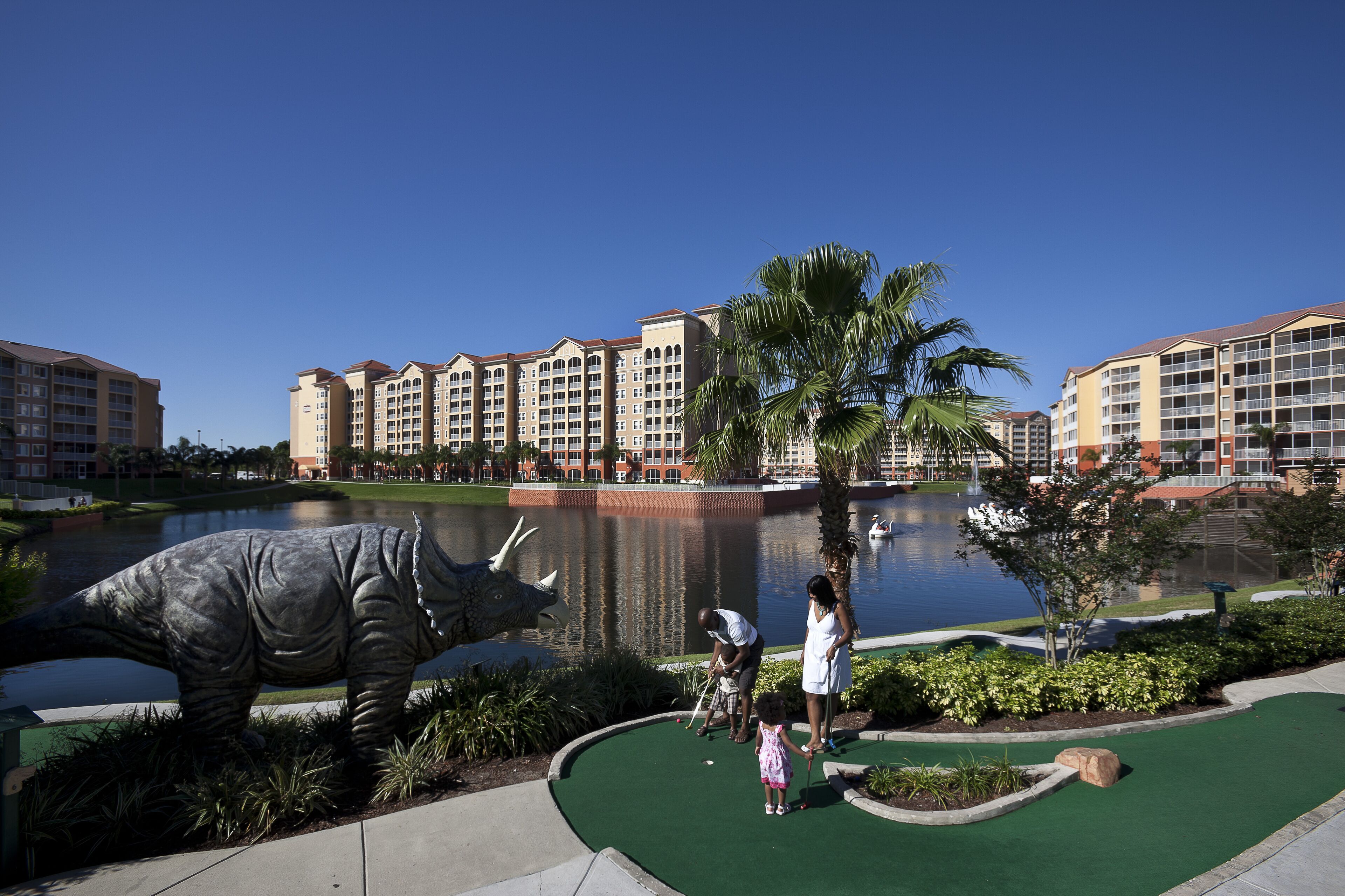 Ein Golfplatz mit Dinosaurier-Statue, Teich und Hotelgebäuden im Hintergrund.