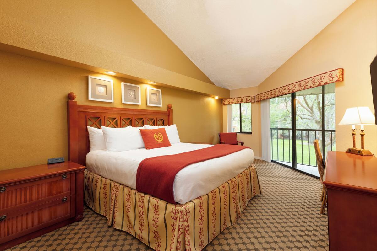 A hotel room with a large bed, a wooden headboard, a nightstand, a desk with a lamp, and a window with a view of greenery.