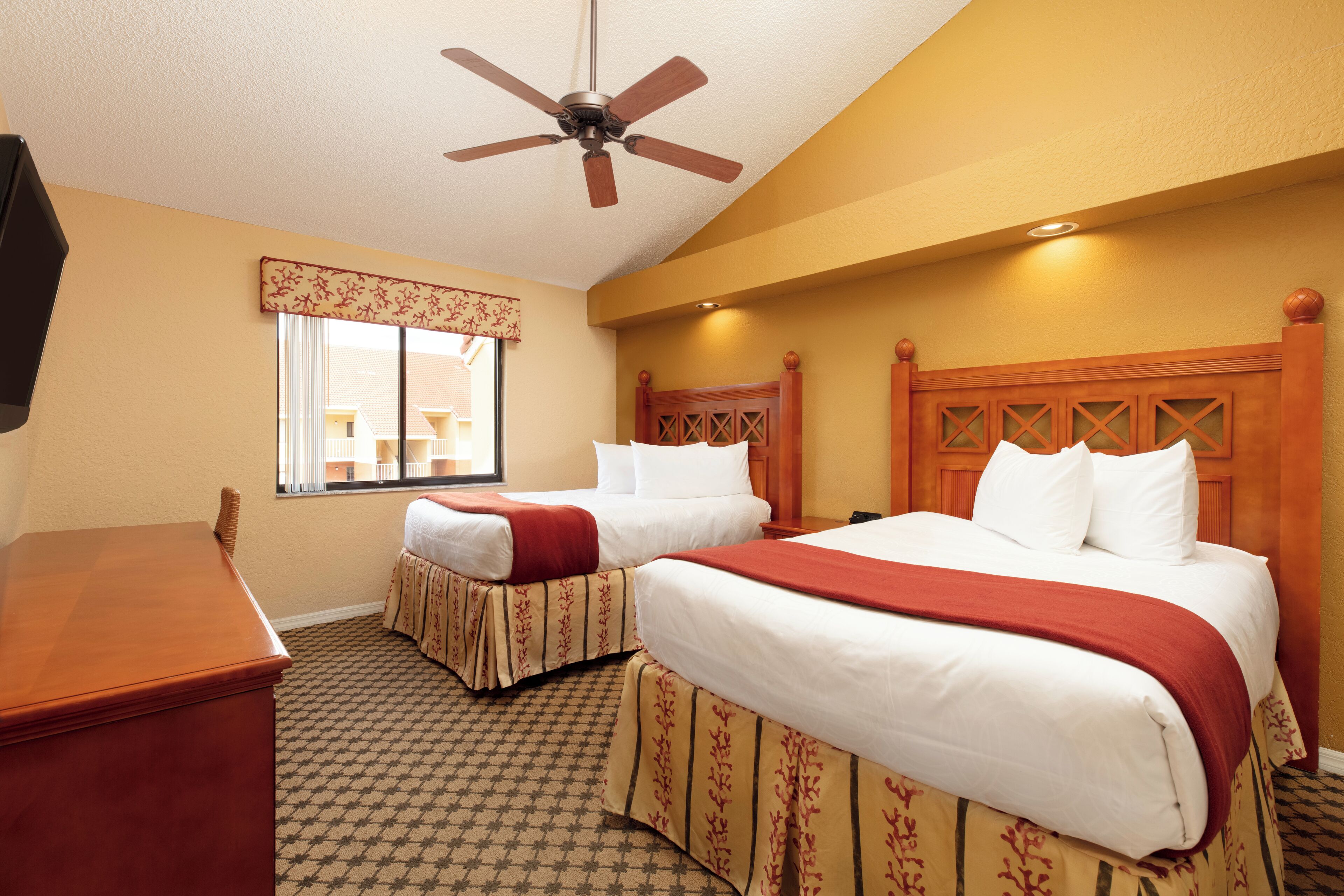 A hotel room with two beds, a ceiling fan, a desk, and a window with a view of the outside.