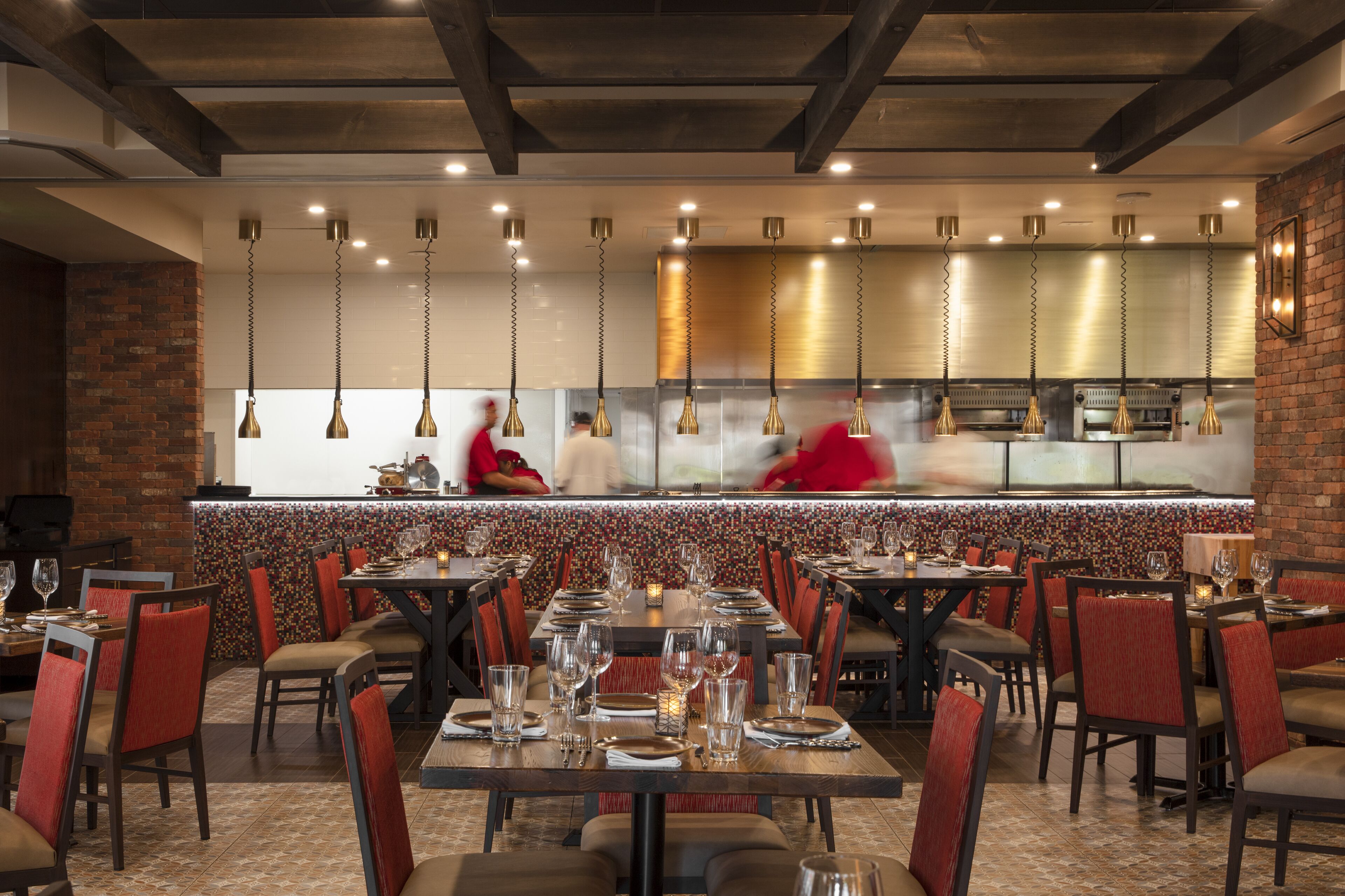 Un restaurante con varias mesas dispuestas para comer, una zona de cocina y lámparas colgantes.