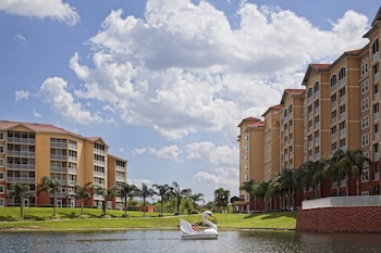 A hotel room with a large bed, a wooden headboard, a nightstand, a desk with a lamp, and a window with a view of greenery. at Westgate Vacation Villas Resort
