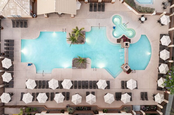 Vista aérea de un área de piscina de hotel con múltiples sillas de descanso y sombrillas, una piscina central y una pequeña cascada.