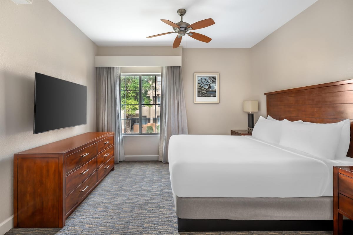 Een hotelkamer met een bed, een televisie, een houten dressoir, een plafondventilator en een raam met gordijnen.