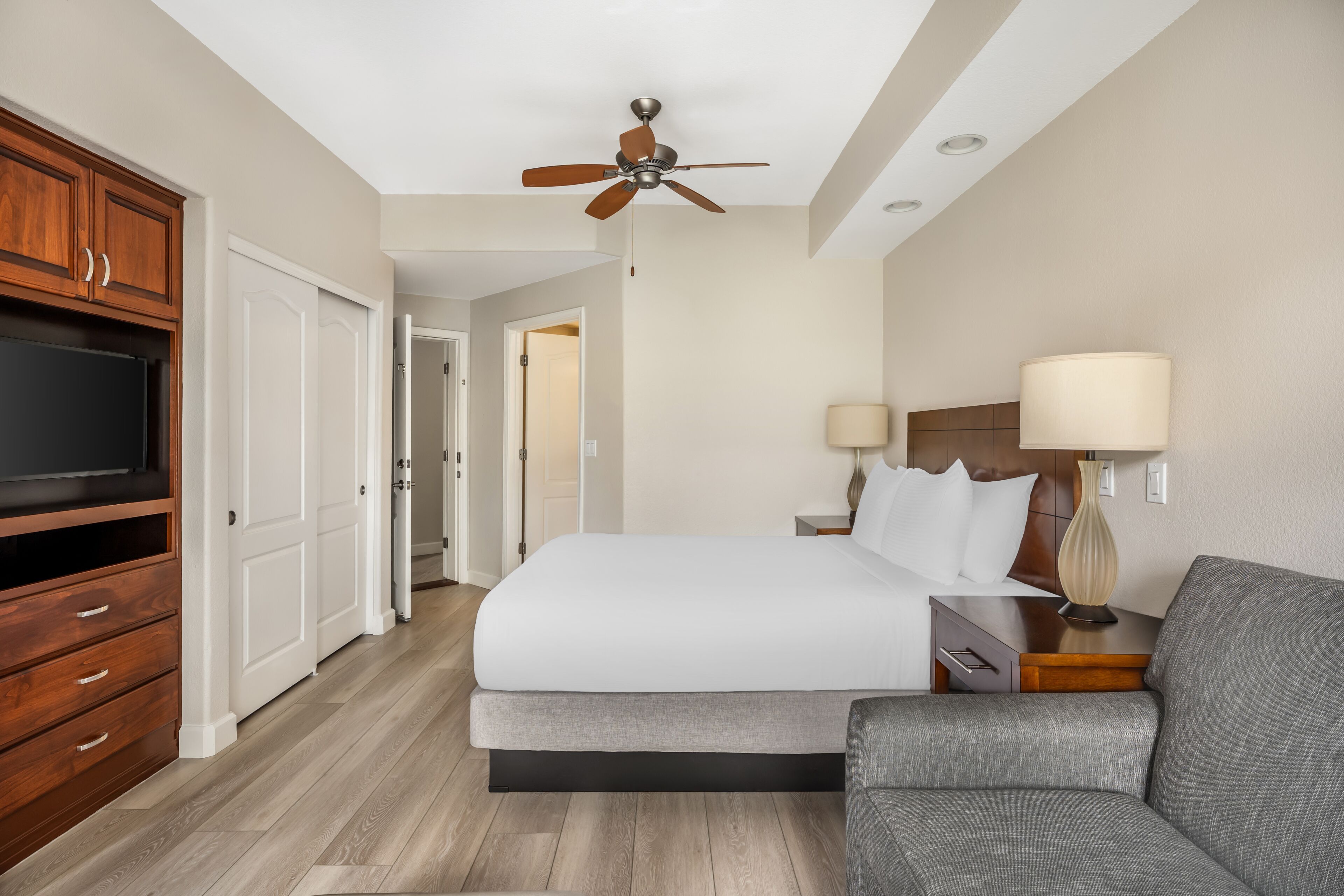 A hotel room with a bed, a TV, a ceiling fan, a nightstand, a lamp, and a sofa.