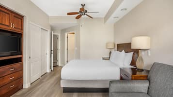 A hotel room with a bed, a TV, a ceiling fan, a nightstand, a lamp, and a sofa.