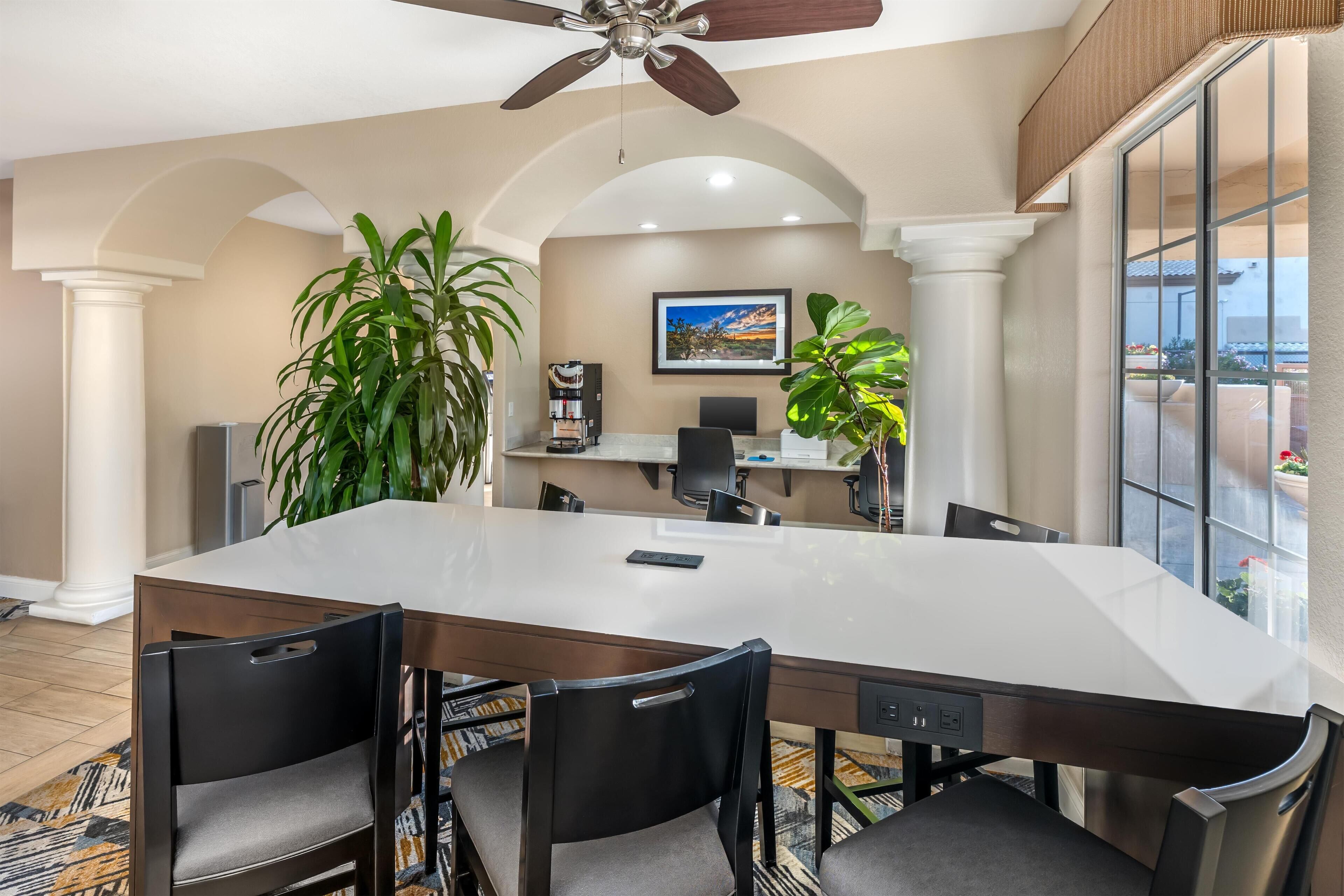 Une salle de conférence avec une grande table blanche, des chaises noires, un ventilateur de plafond et des plantes en pot.