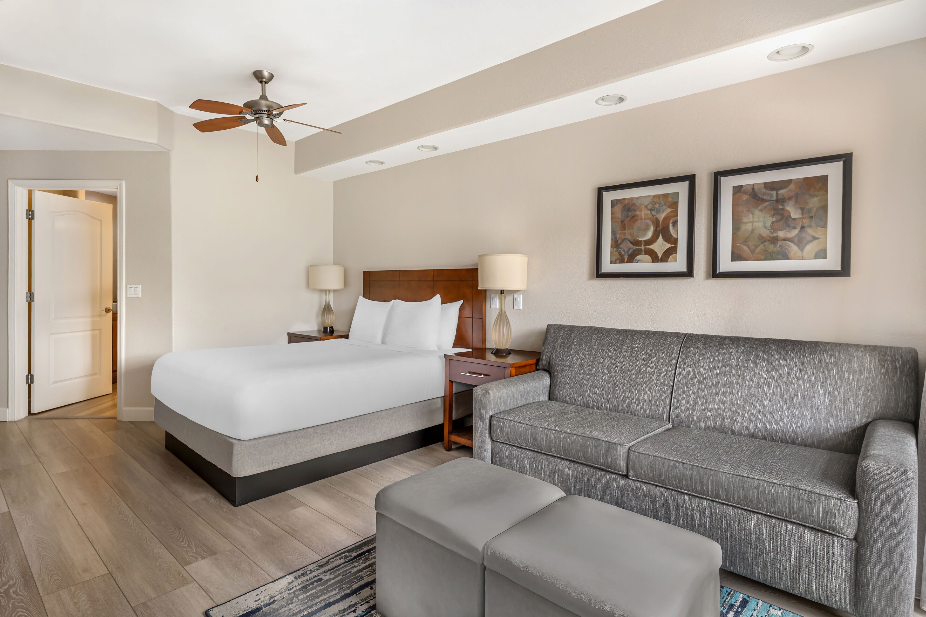 A hotel room with a bed, a sofa, a ceiling fan, two bedside lamps, and two framed pictures on the wall.