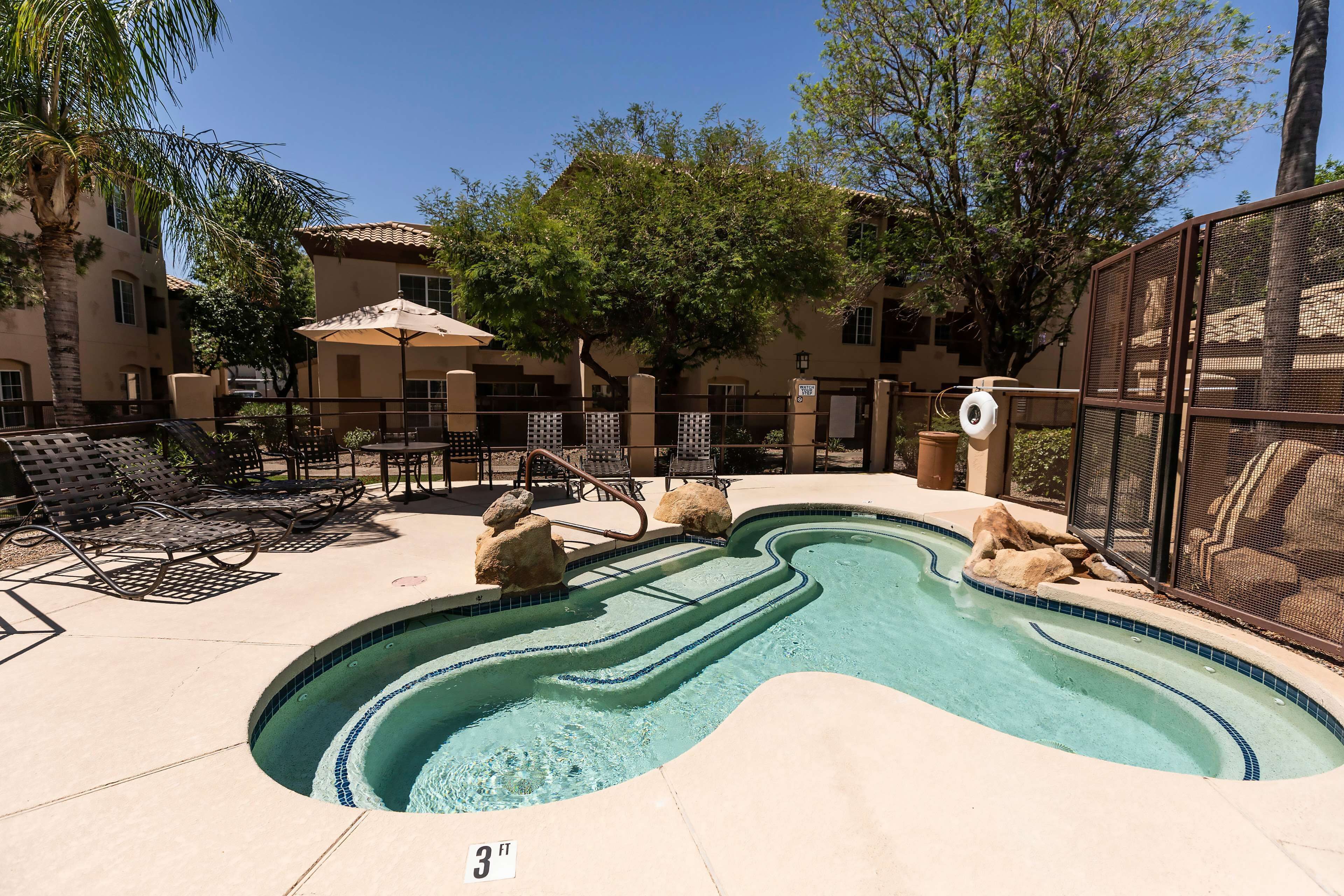 A swimming pool with a 3 ft depth marker, surrounded by lounge chairs and umbrellas, with a building and trees in the background.