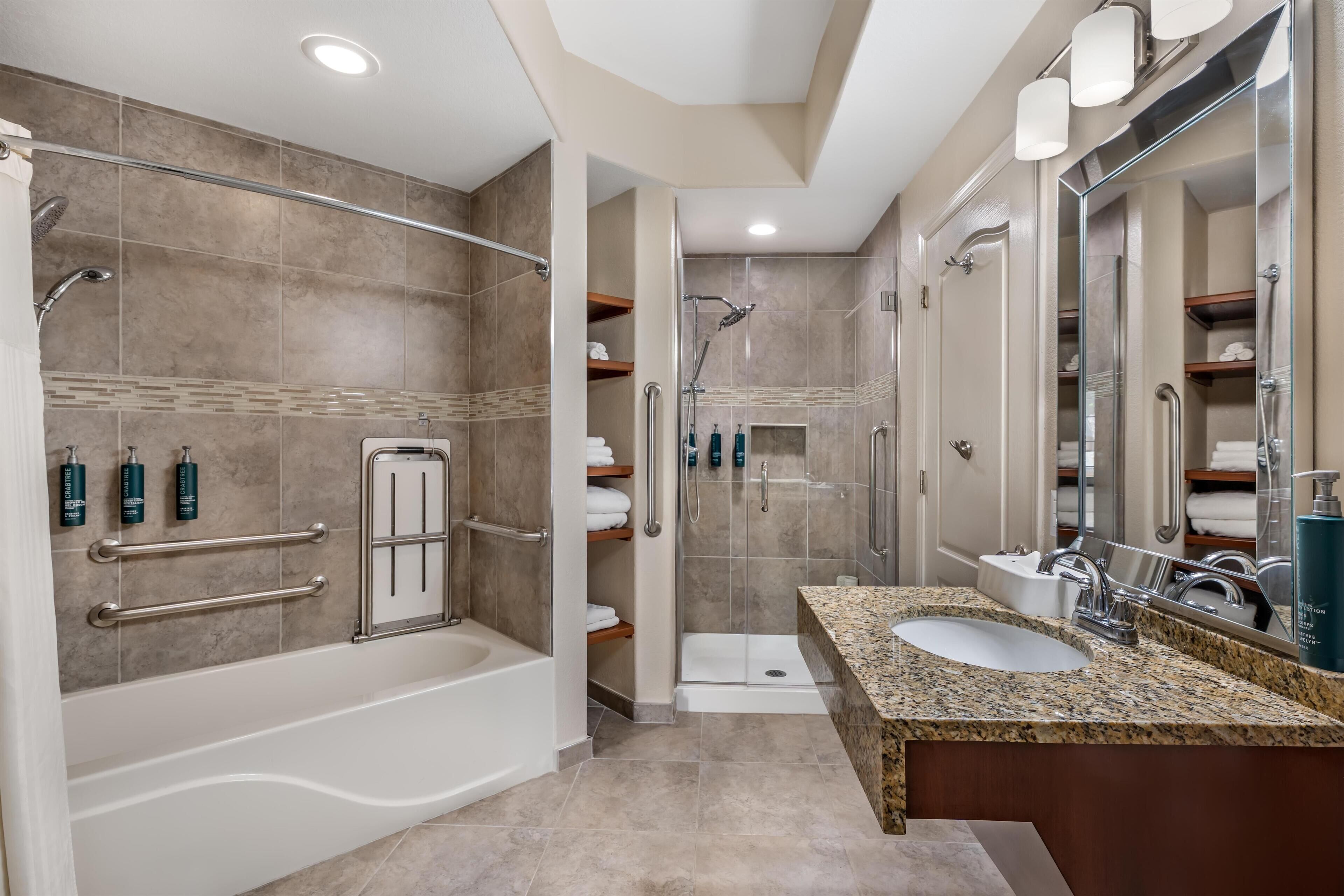 A modern bathroom with a bathtub, shower, and sink, featuring grab bars, shelves with toiletries, and a large mirror.