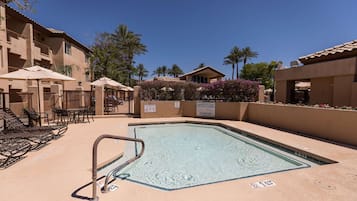 A pool area with lounge chairs and umbrellas, surrounded by buildings with balconies and palm trees.
