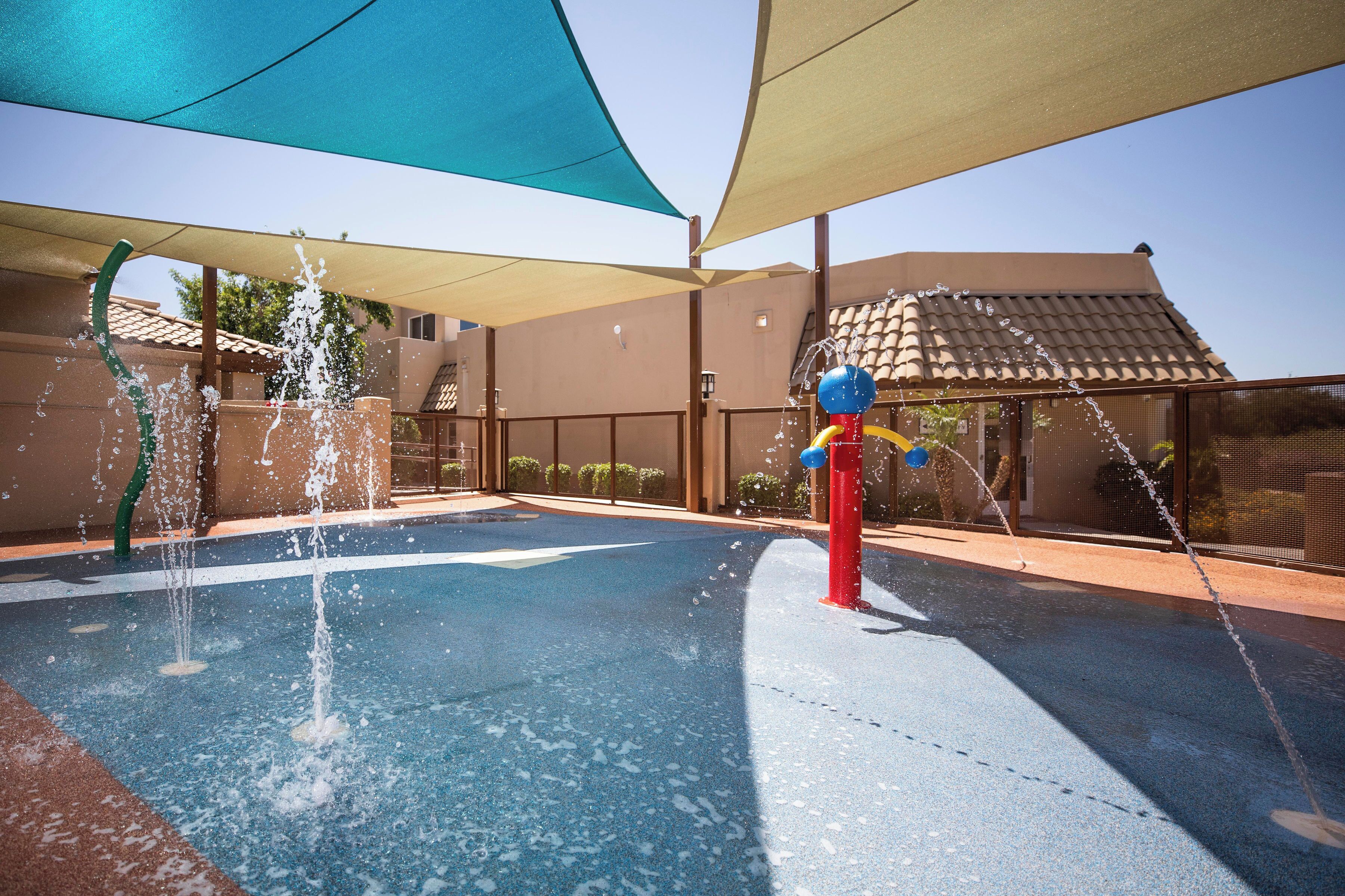 Zona de juegos acuáticos al aire libre con múltiples chorros de agua y un área de descanso cubierta.