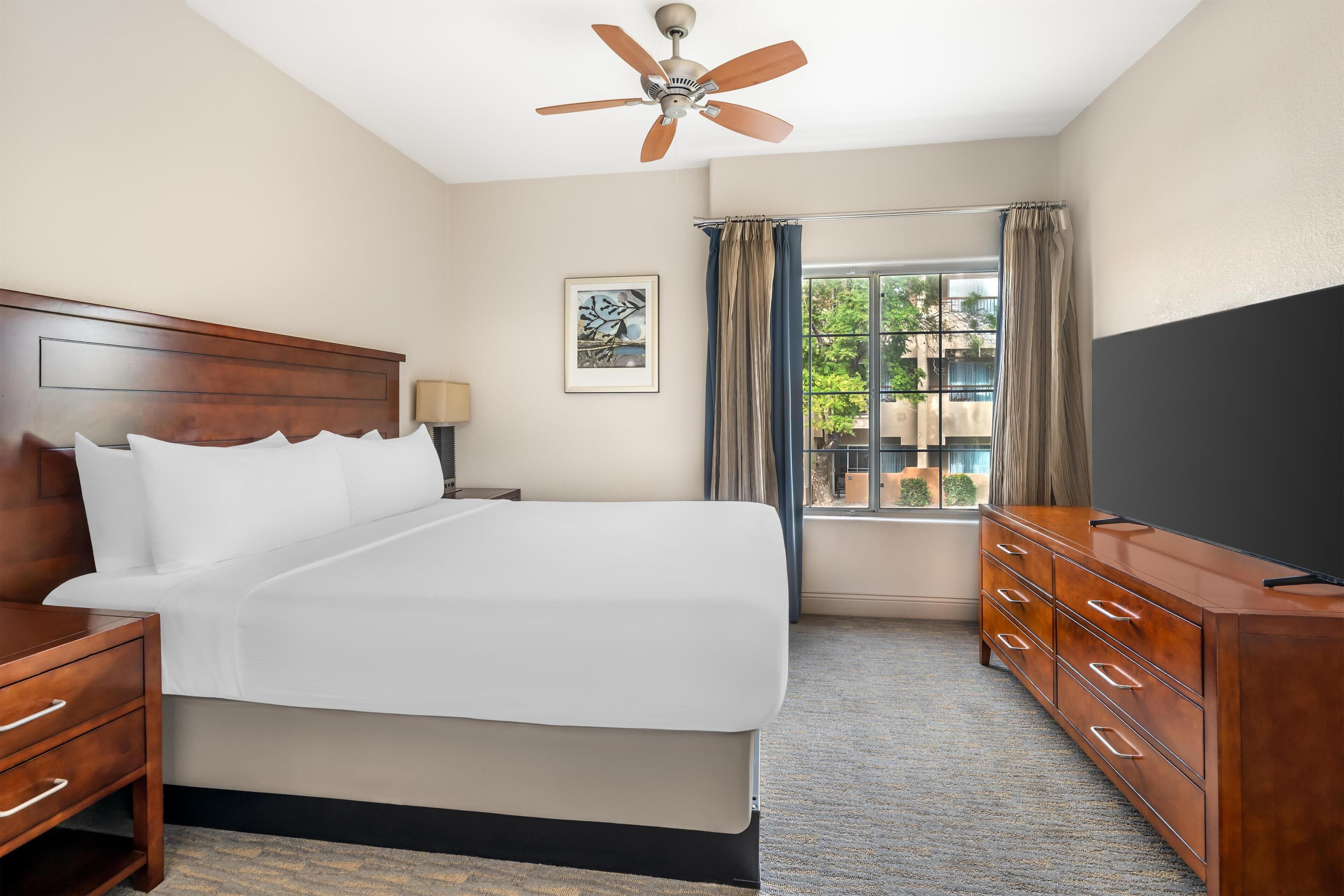 A hotel room with a large bed, wooden headboard, a dresser, a ceiling fan, a window with curtains, and a TV.