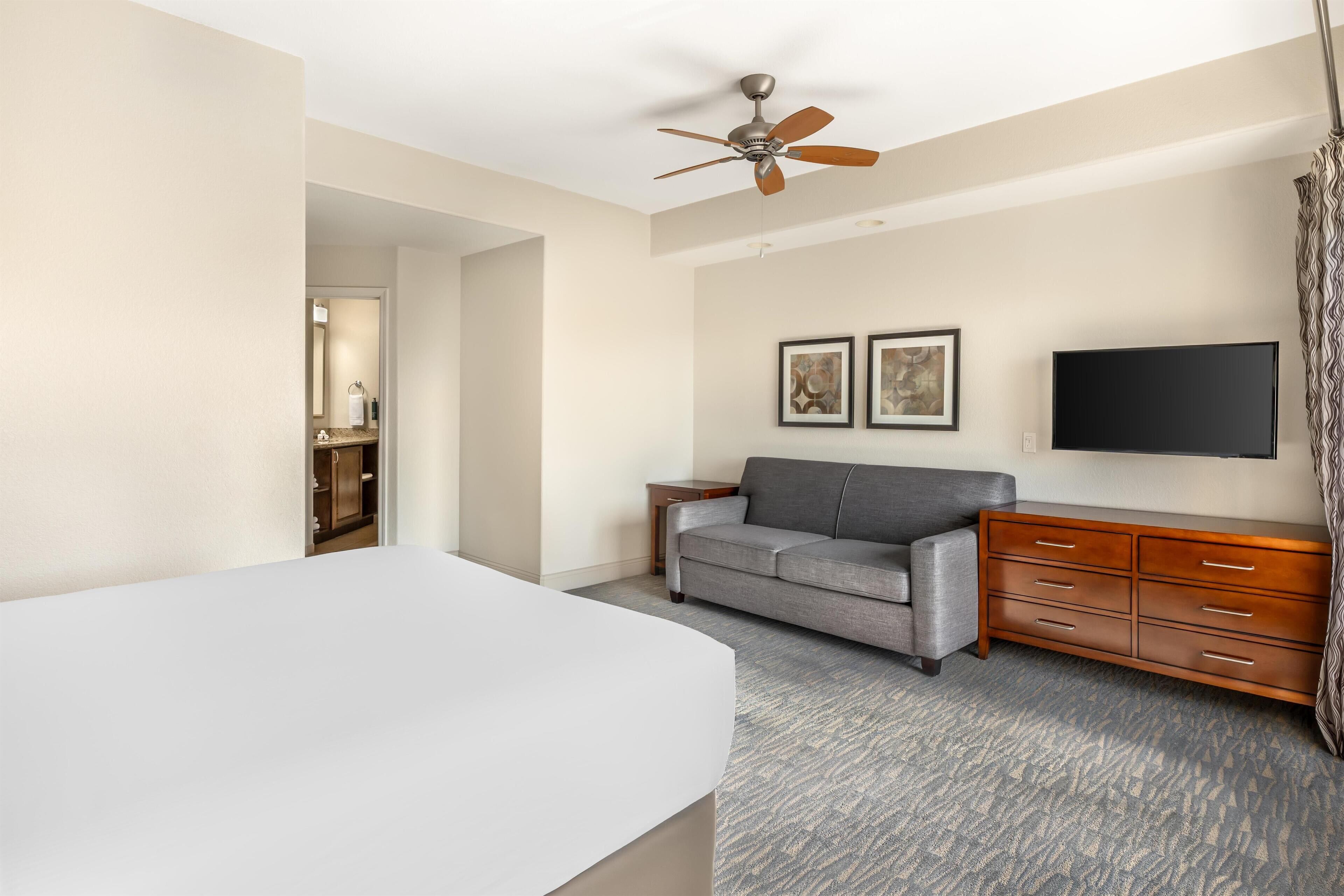 A hotel room with a bed, a gray sofa, a wooden dresser, a ceiling fan, and a flat-screen TV mounted on the wall.