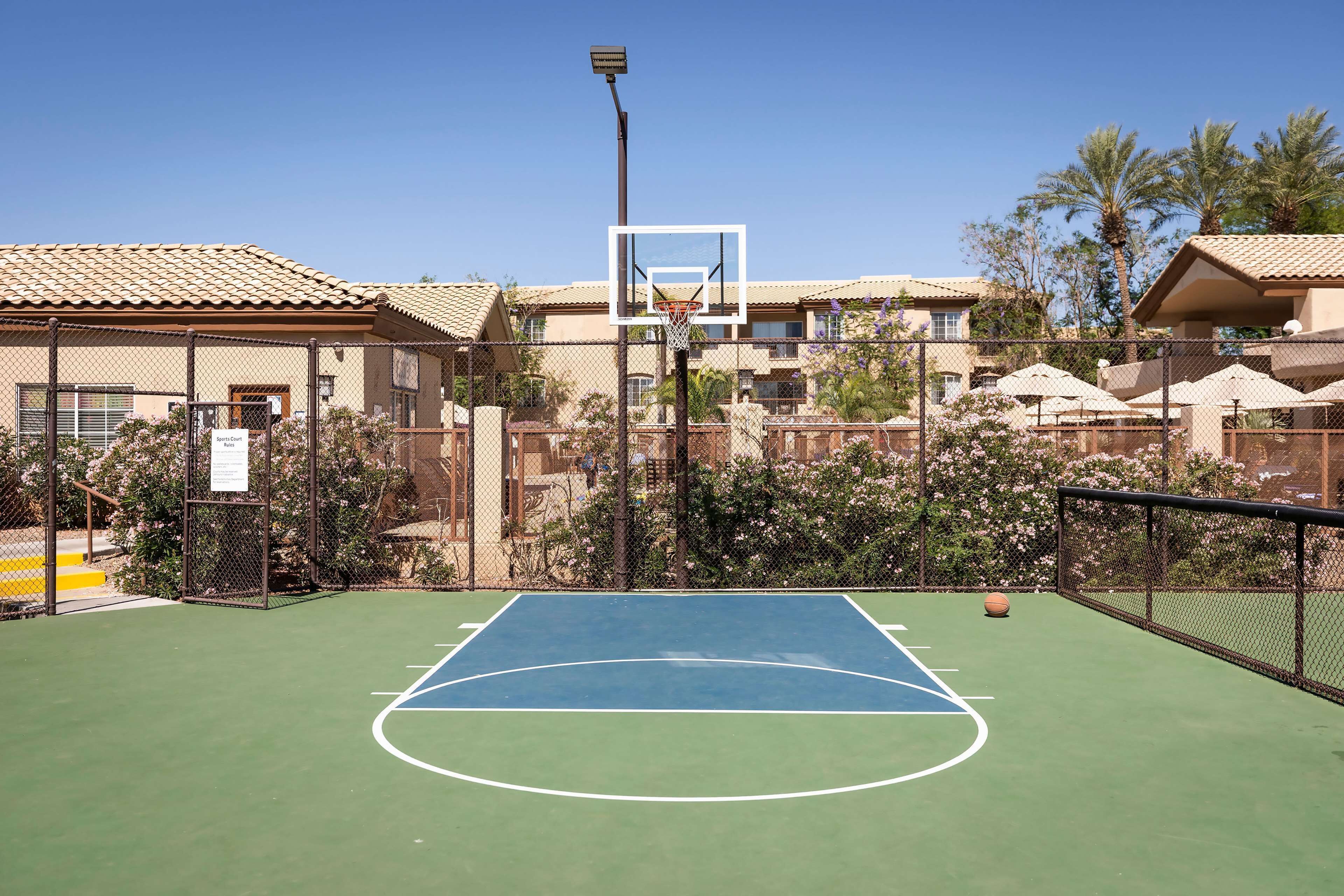 Una cancha de baloncesto al aire libre rodeada por una cerca de malla metálica, con un aro de baloncesto en el centro.