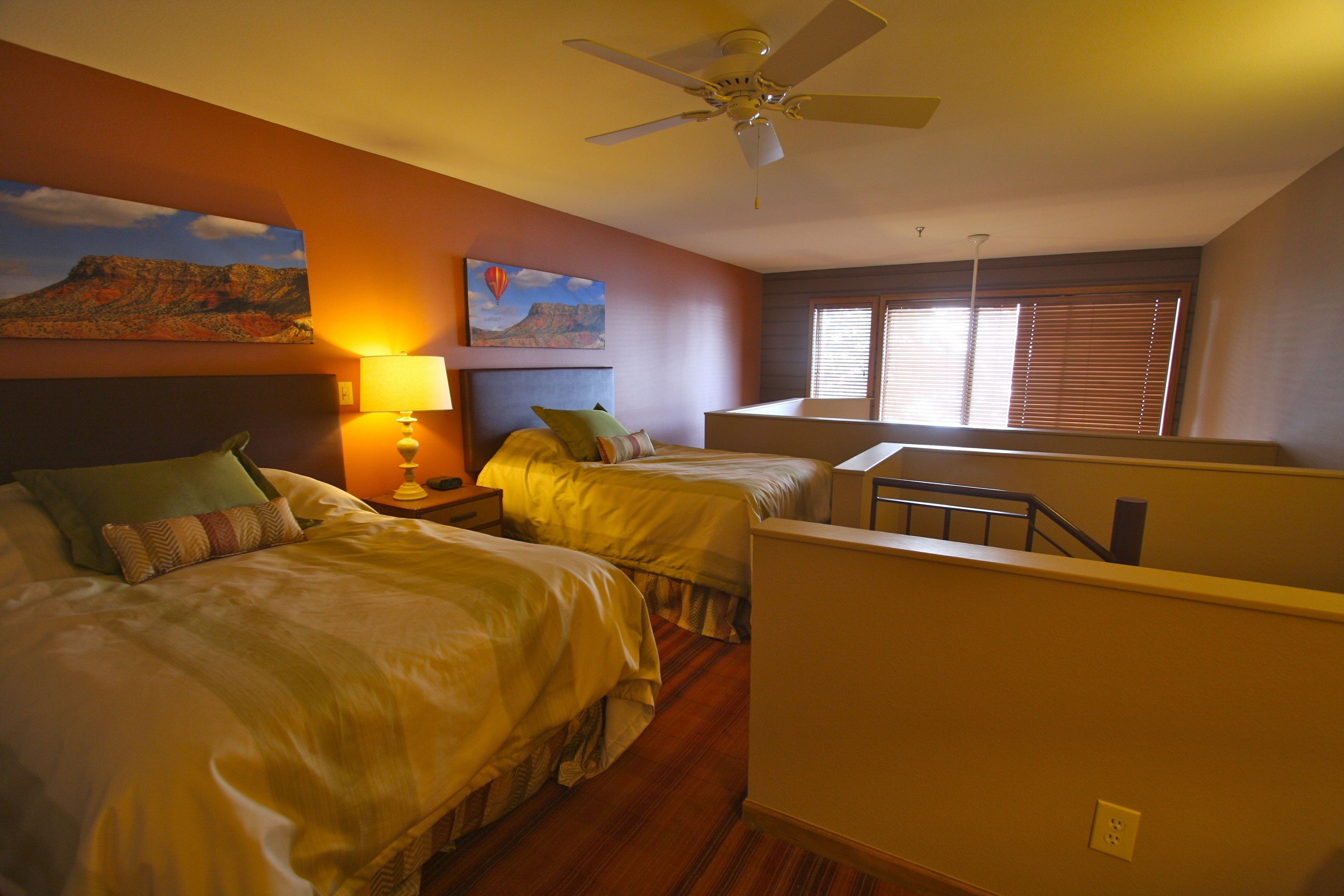 Une chambre d’hôtel avec deux lits, un ventilateur de plafond, un tableau représentant une montagne et une fenêtre avec des stores.