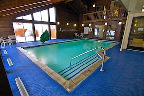 Une piscine intérieure avec un sol carrelé de bleu, une rambarde métallique et un balcon en bois donnant sur la piscine.