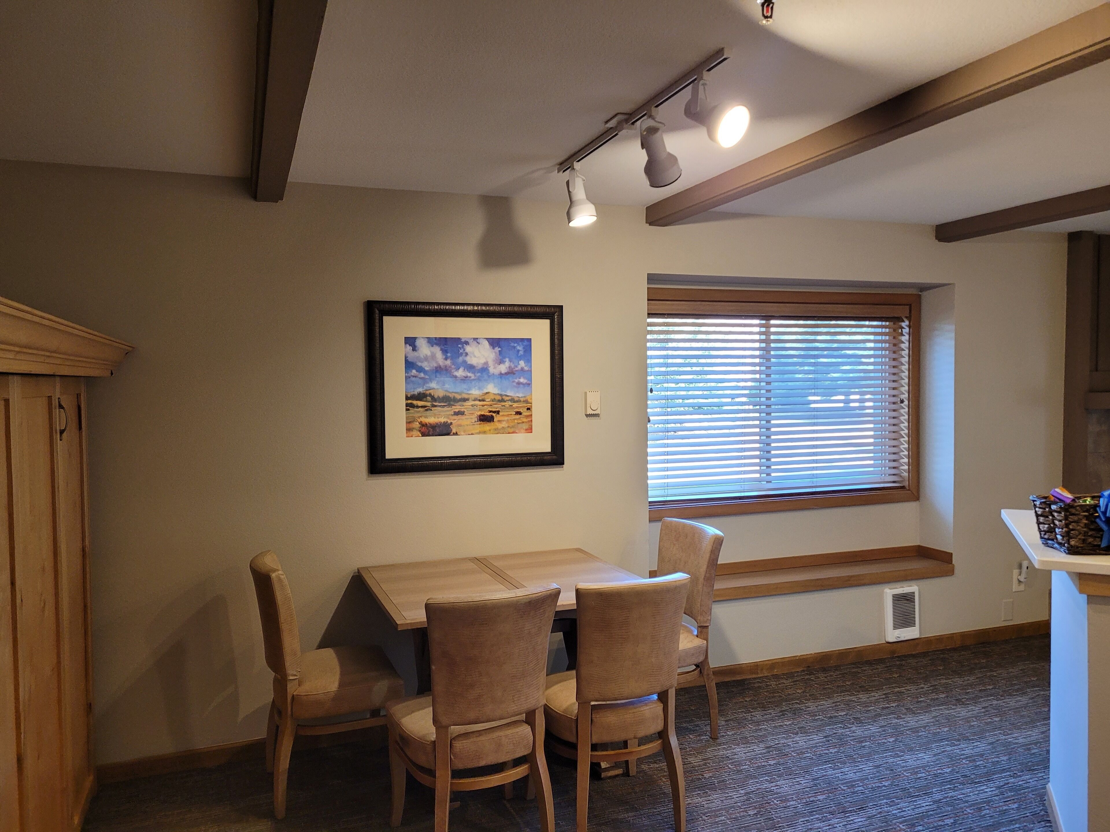 A dining area with a wooden table and chairs, a framed landscape painting on the wall, a window with blinds, and a ceiling light fixture.