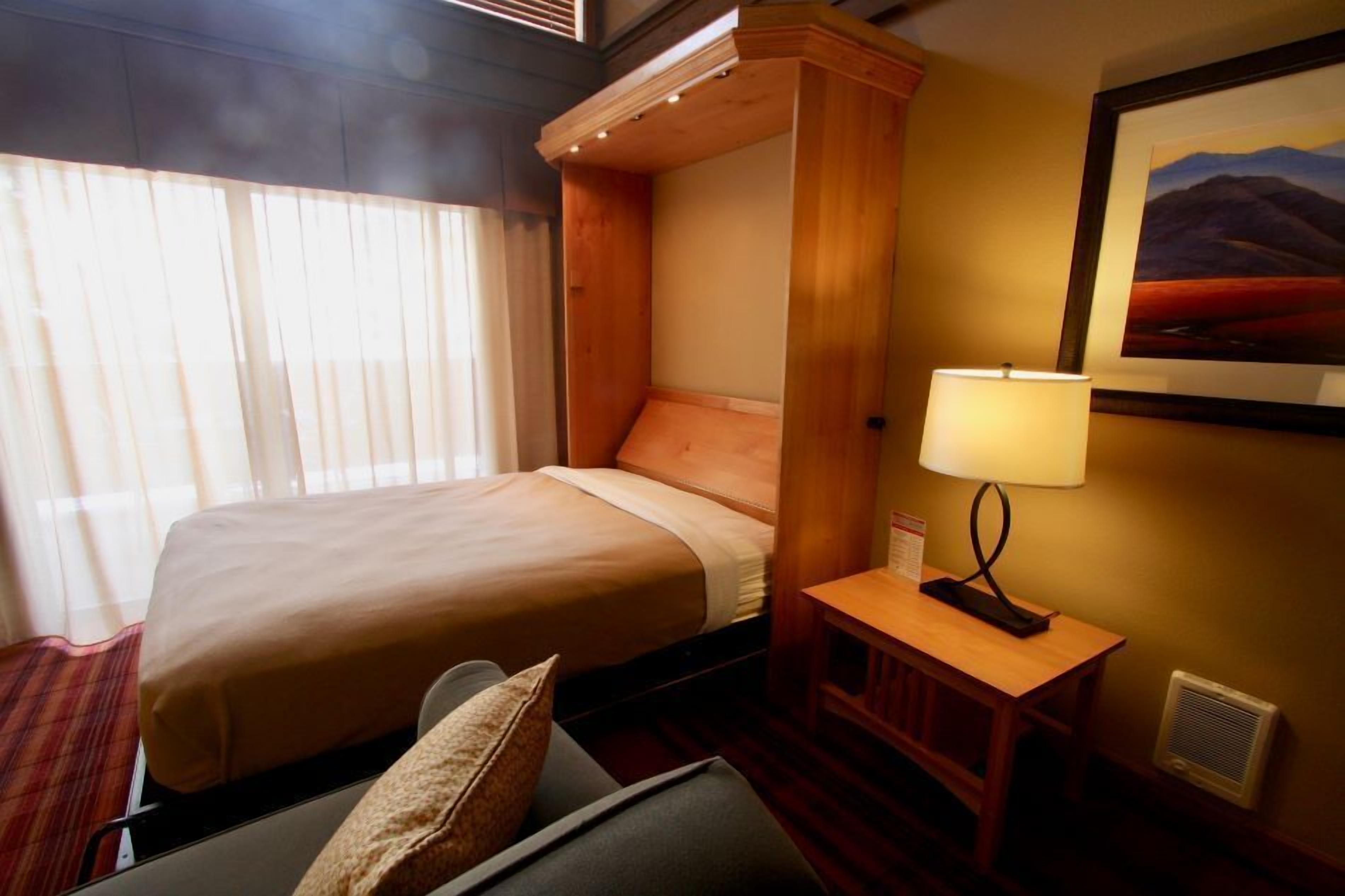 A hotel room with a large bed, a bedside table with a lamp, a window with curtains, and a framed picture on the wall.