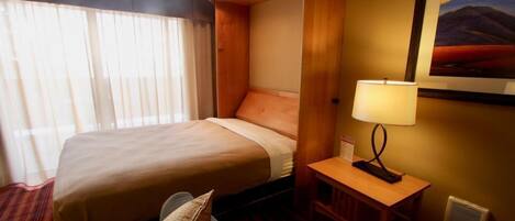 A hotel room with a large bed, a bedside table with a lamp, a window with curtains, and a framed picture on the wall.