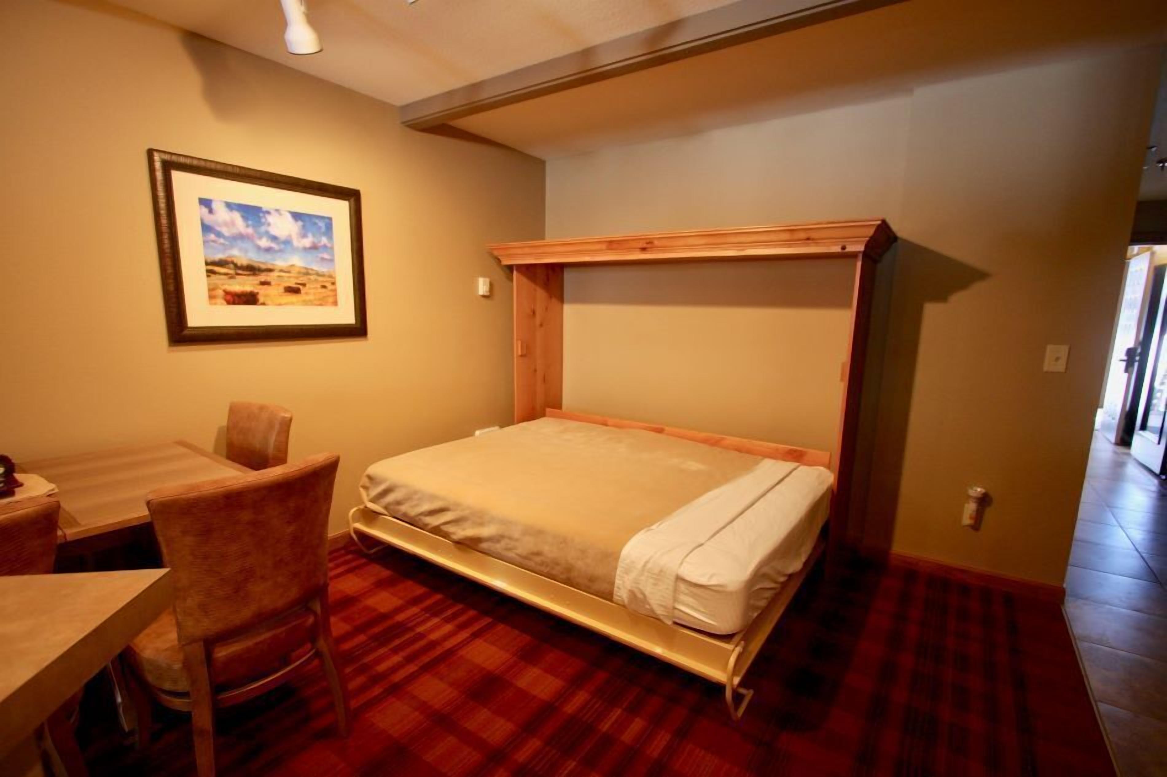 A room with a bed, a desk, and a chair. There is a framed picture on the wall.