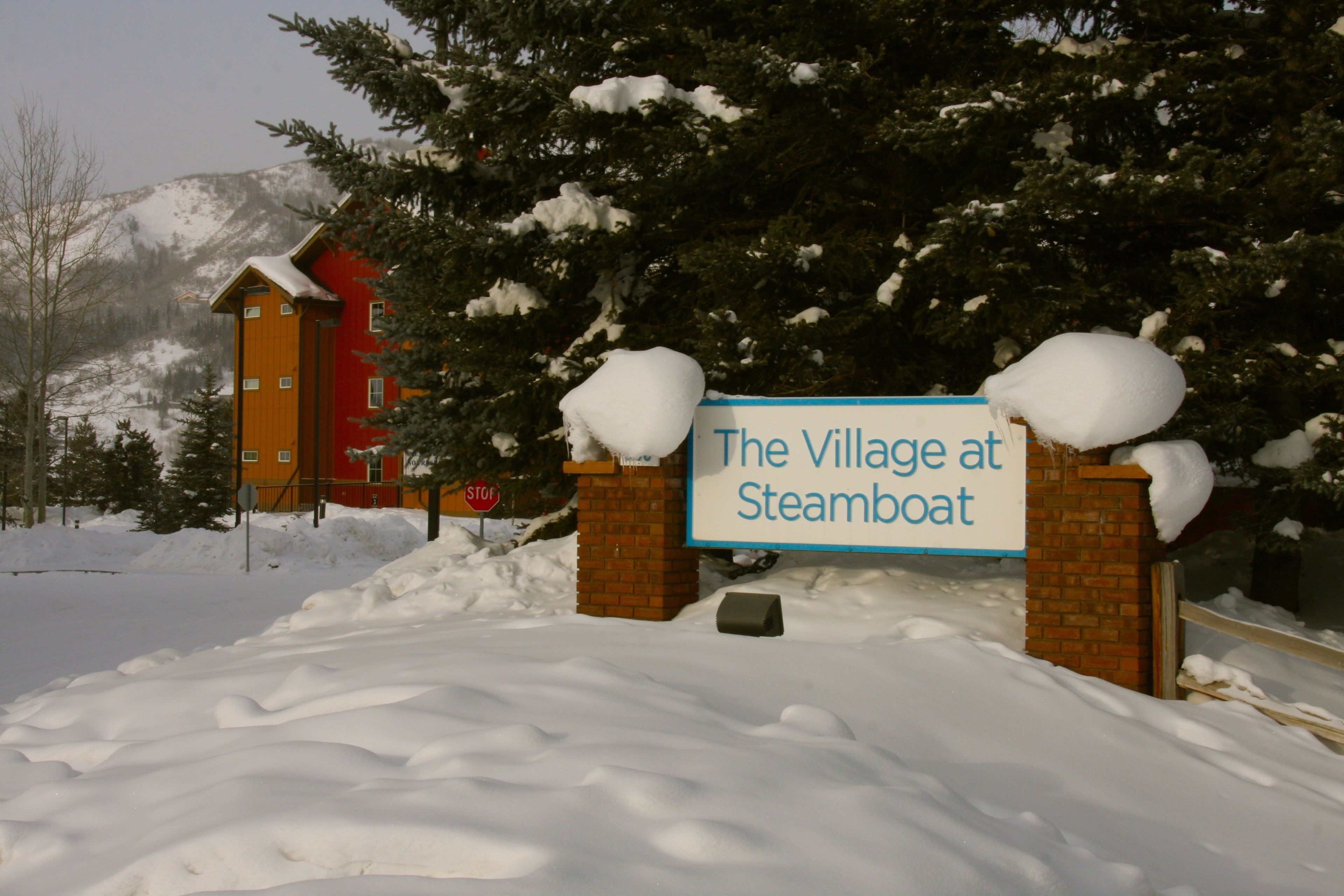Een bord bedekt met sneeuw met de tekst 'The Village at Steamboat', met een rood gebouw op de achtergrond.