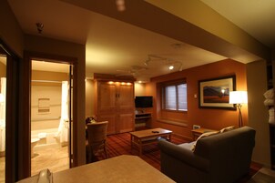 Habitación de hotel con piso de madera, un sofá, un televisor, un cuadro y una ventana con persianas.