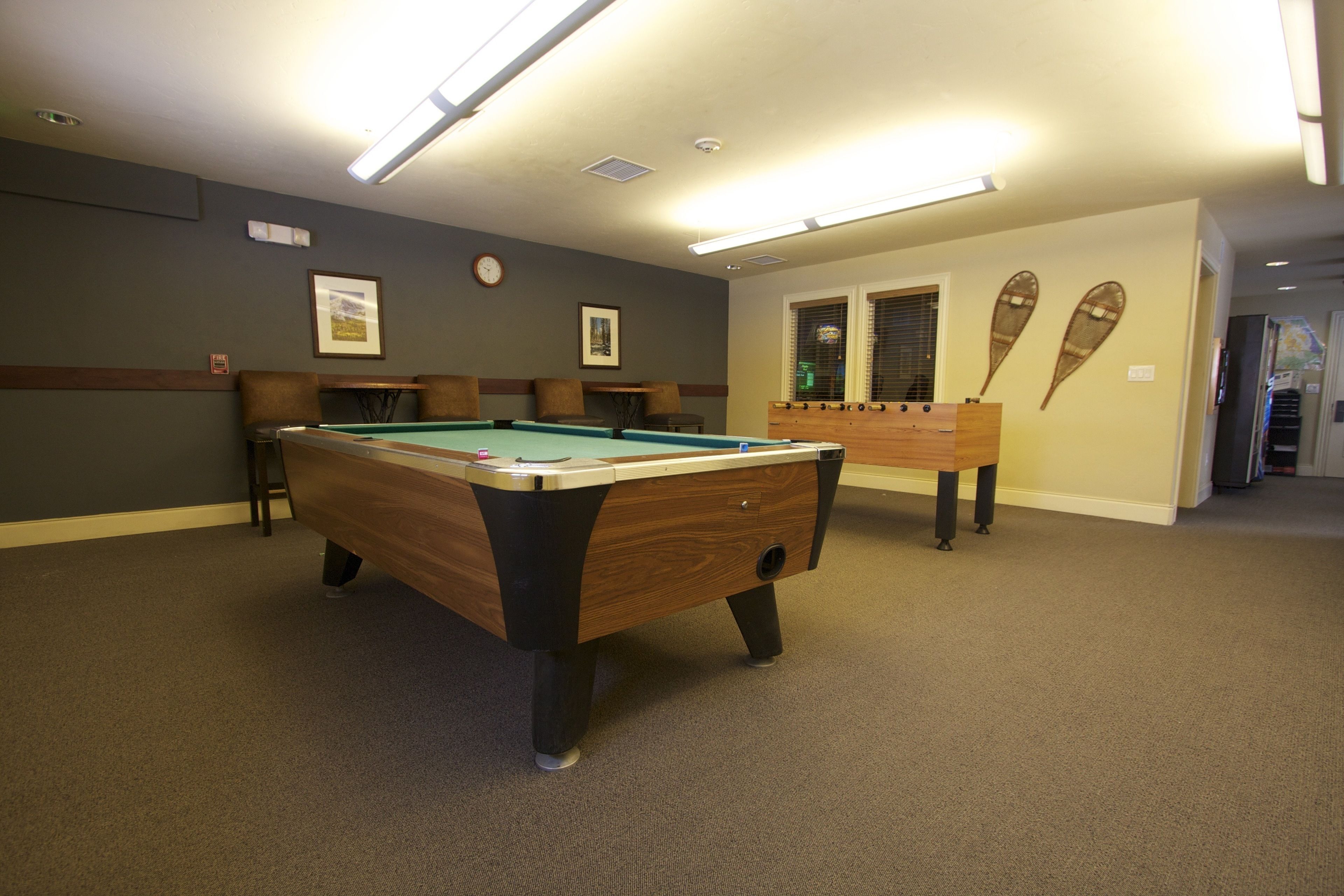 A pool table in a room with a carpeted floor, framed pictures on the wall, and a window with blinds.