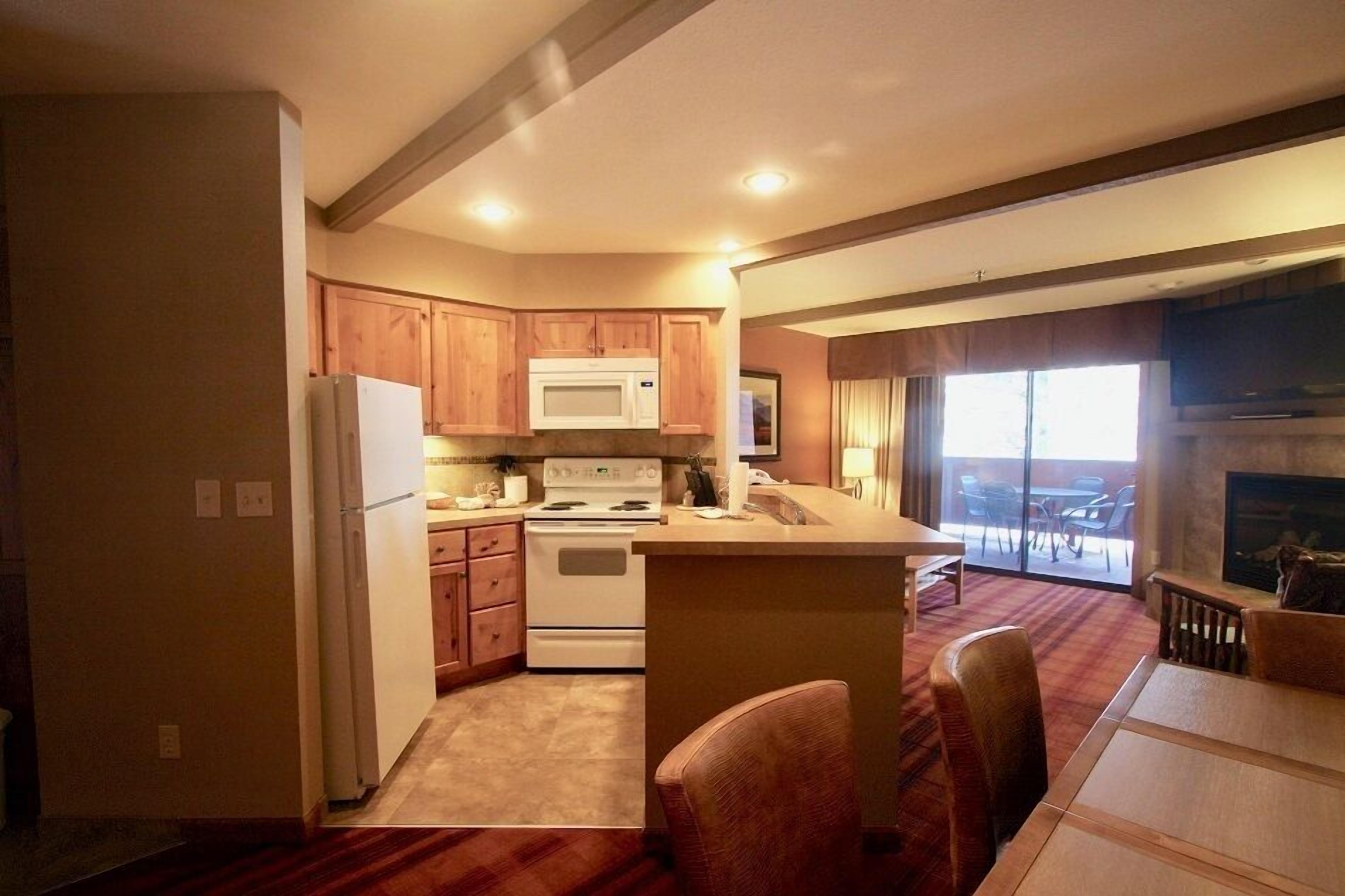 A compact kitchen with wooden cabinets, a microwave, stove, and a refrigerator. There is a dining area with a table and chairs, and a fireplace in the living room.