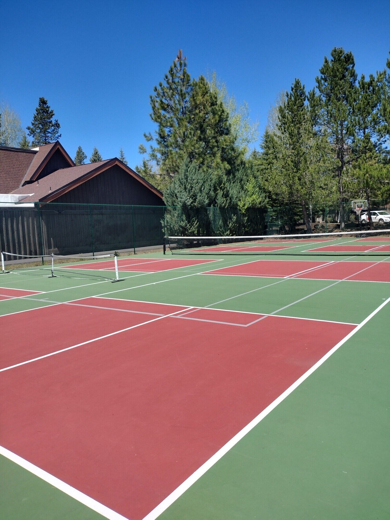 An outdoor tennis court with a red and green surface, surrounded by trees and a building with a sloped roof.