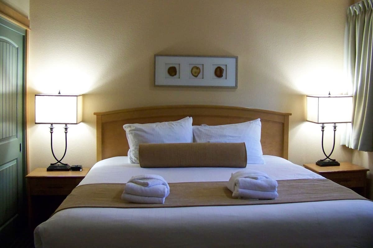 Une chambre d’hôtel avec un grand lit, deux lampes de chevet, une tête de lit en bois et un tableau encadré au-dessus du lit.