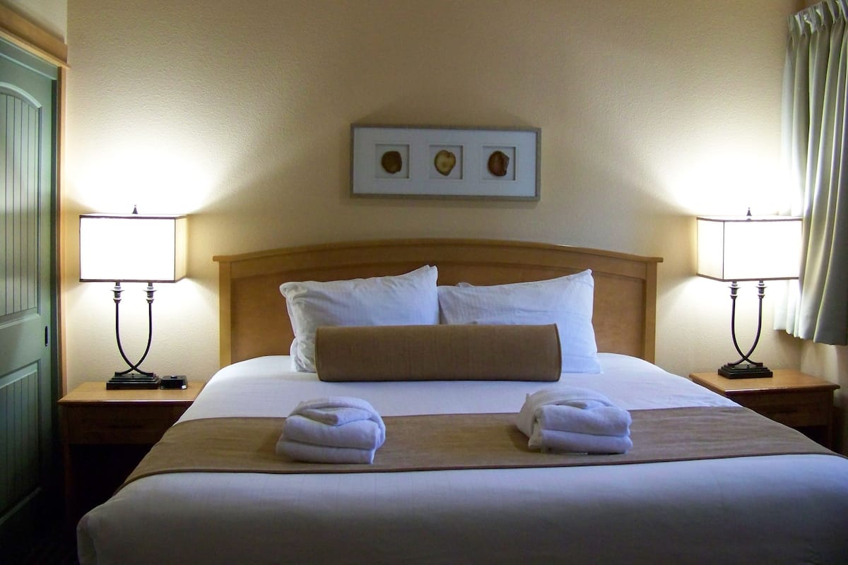 Une chambre d’hôtel avec un lit, deux lampes de chevet, une tête de lit en bois, un tableau encadré au-dessus du lit et deux serviettes sur le lit.