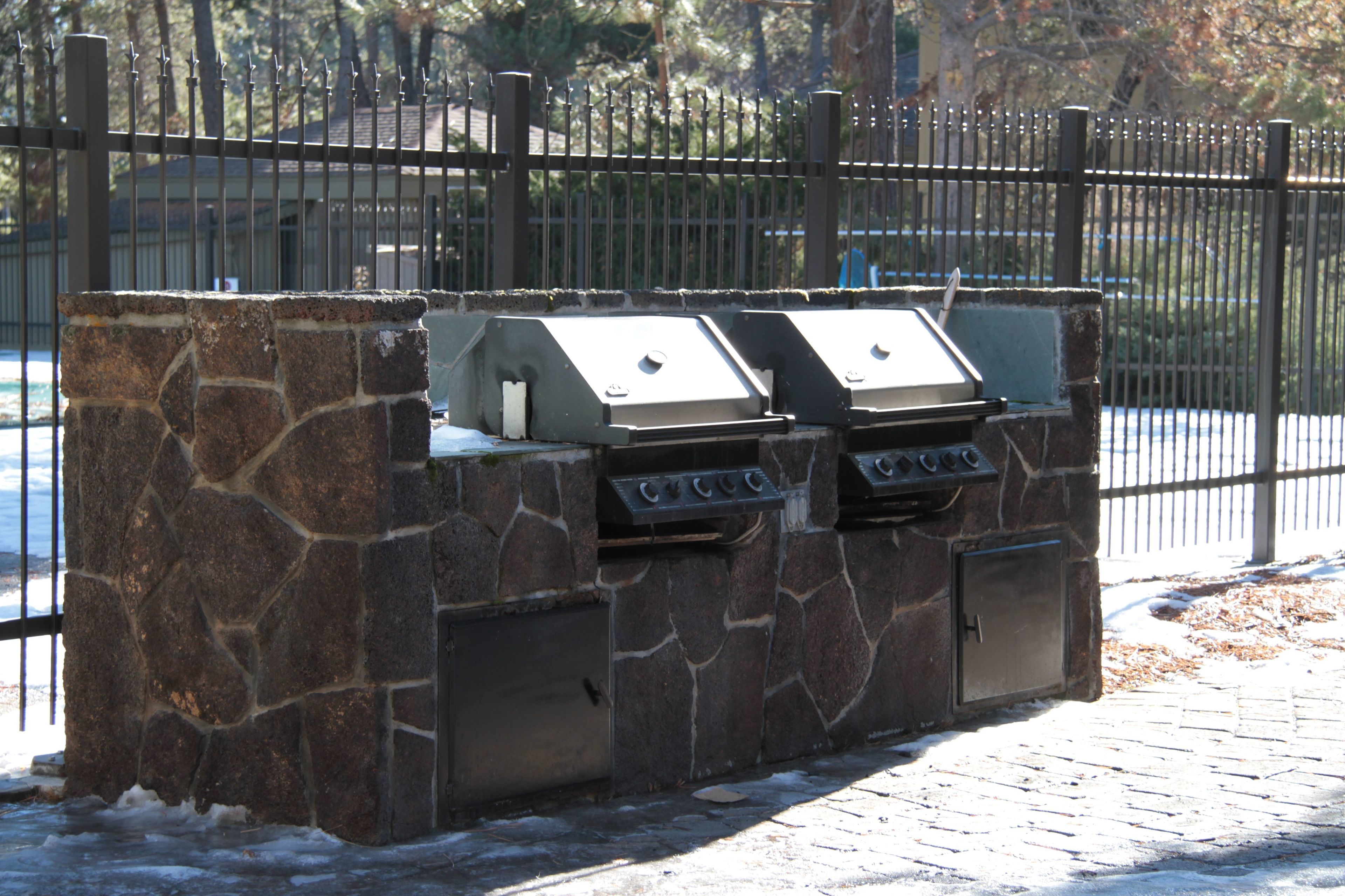 A stone barbecue grill with two burners, located outdoors with a metal fence in the background.