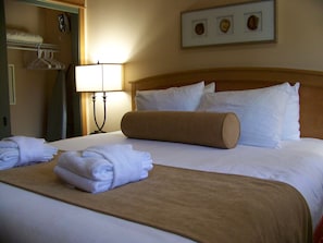 A neatly made bed with white linens, a brown throw blanket, and a folded white towel. There is a bedside lamp, a framed picture on the wall, and a coat rack with hangers.