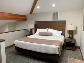 Une chambre d’hôtel avec un grand lit, deux tables de chevet avec des lampes, une tête de lit en bois, une fenêtre avec des stores et un sol recouvert de moquette.